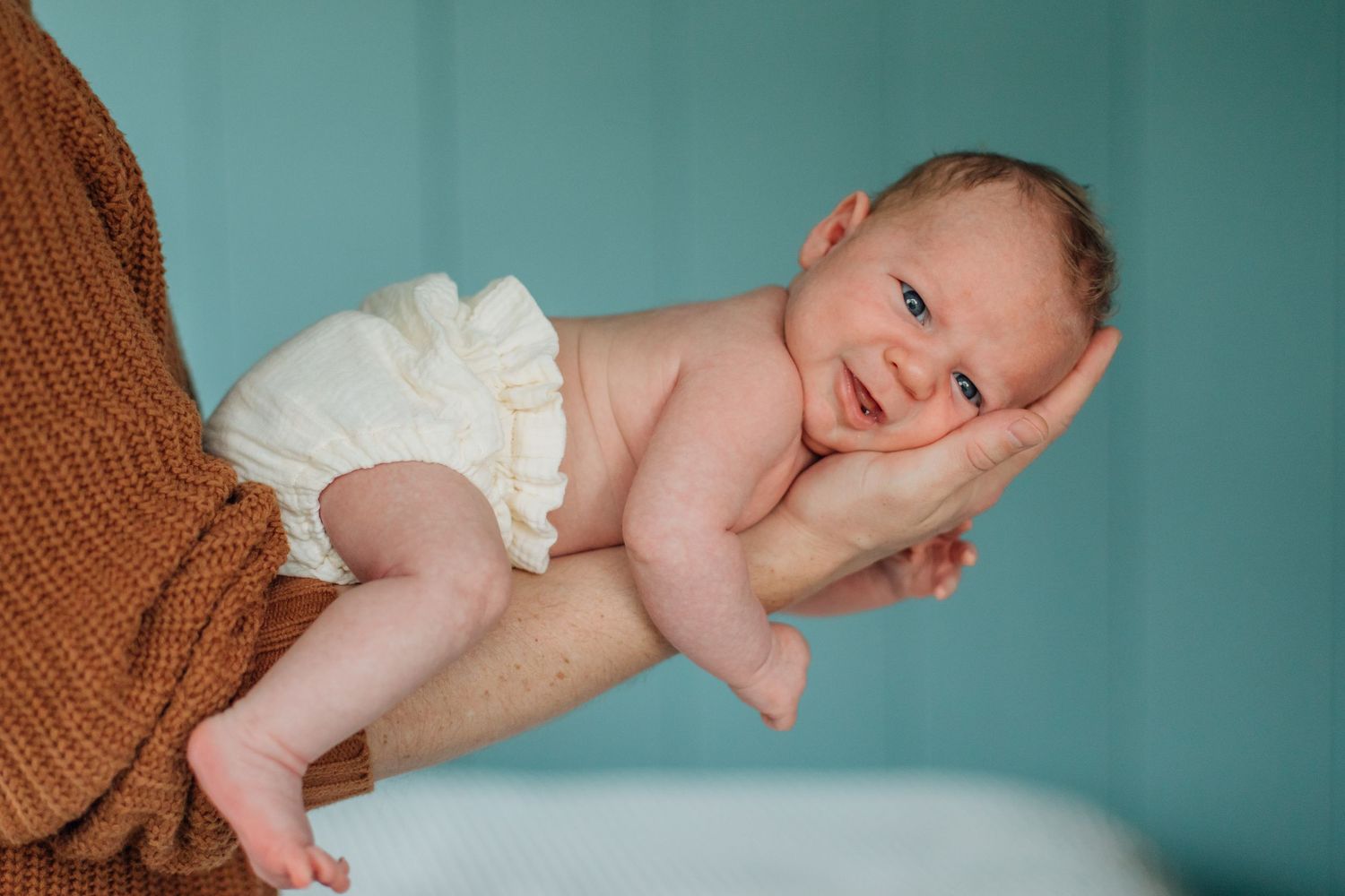 Smiling newborn baby in white diaper held securely on adult's forearm against teal background.