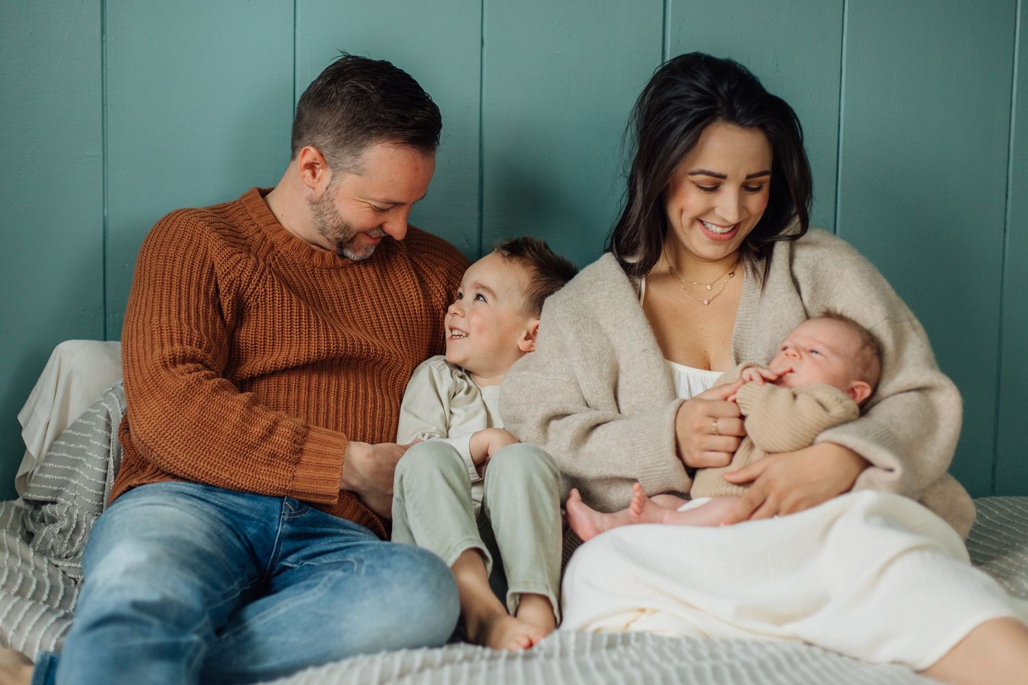 A loving family sits together on a bed, parents smiling as their toddler and newborn bond.