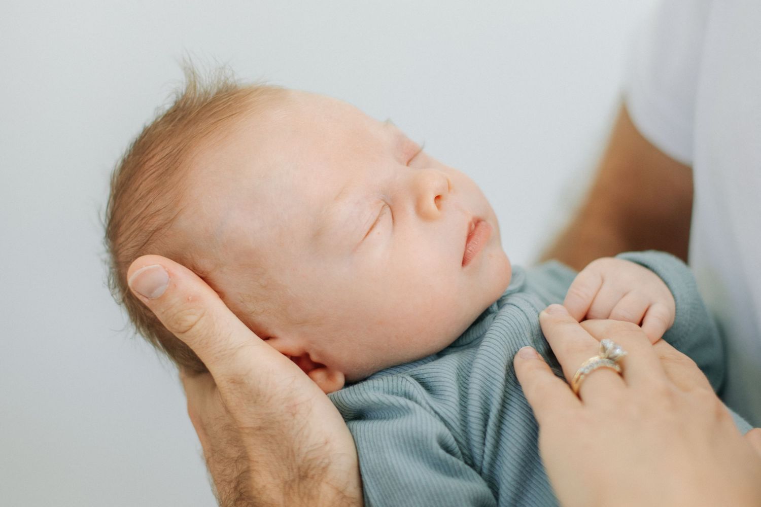 Close-up of peaceful sleeping newborn baby in sage green outfit with parents hands visible.