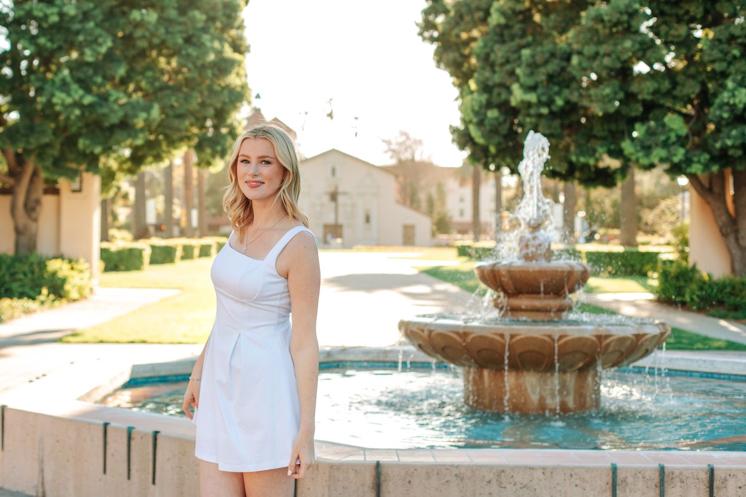 A young blonde woman in a white dress stands beside an ornate stone fountain outdoors.