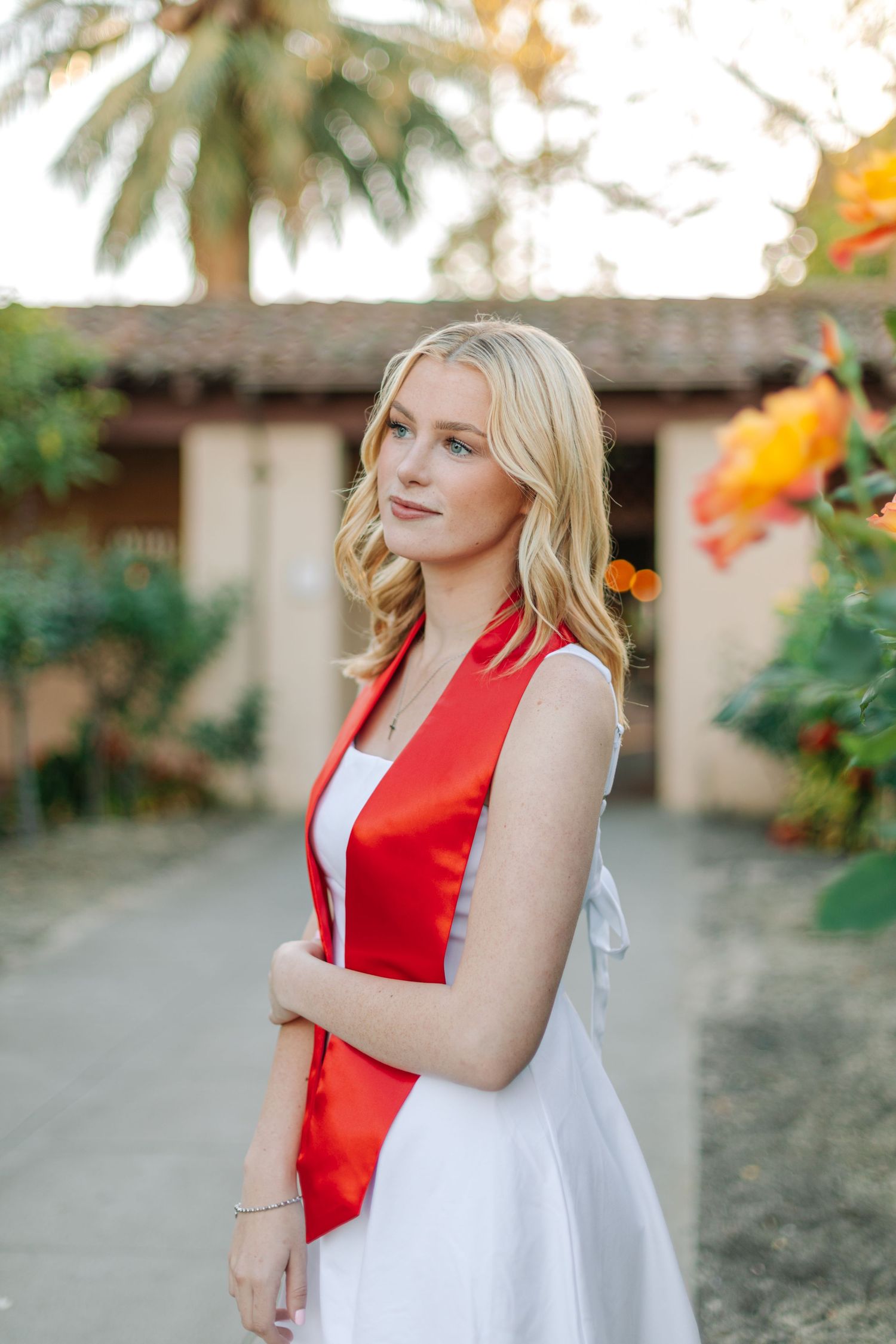 Blonde graduate in white dress and red stole stands in garden with tropical flowers at sunset.