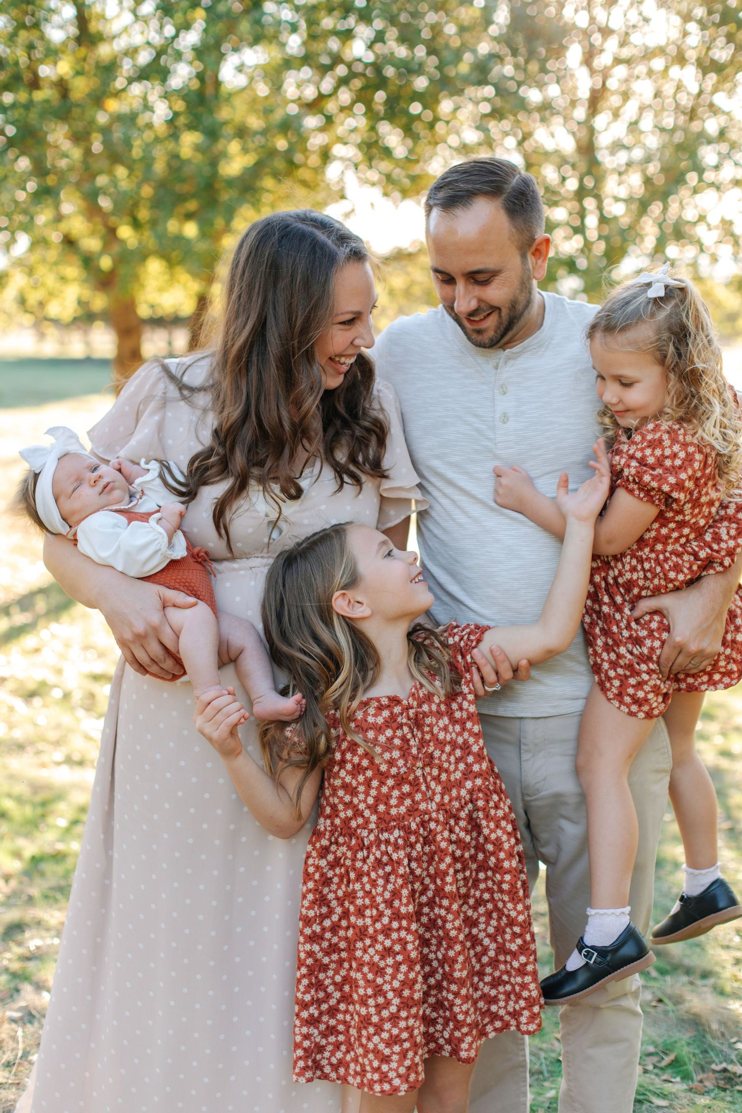 A smiling family of five with three daughters, including a newborn, enjoys a sunny outdoor moment.