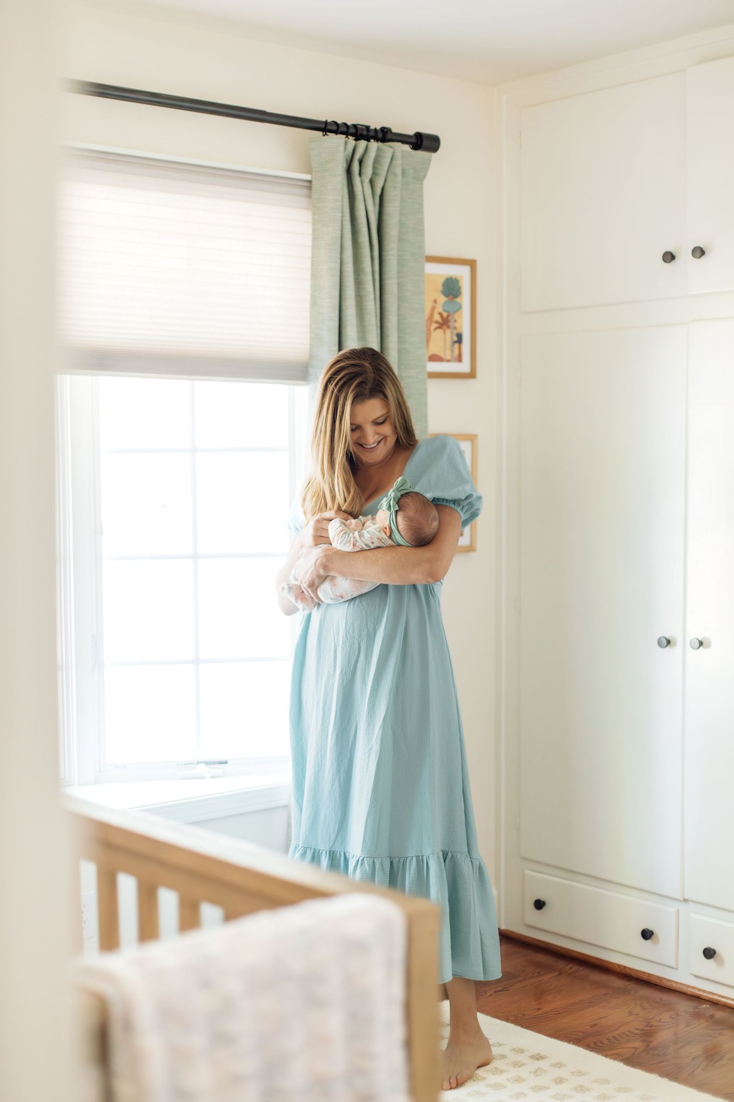 New mother in light blue dress tenderly holding newborn baby by bright nursery window.