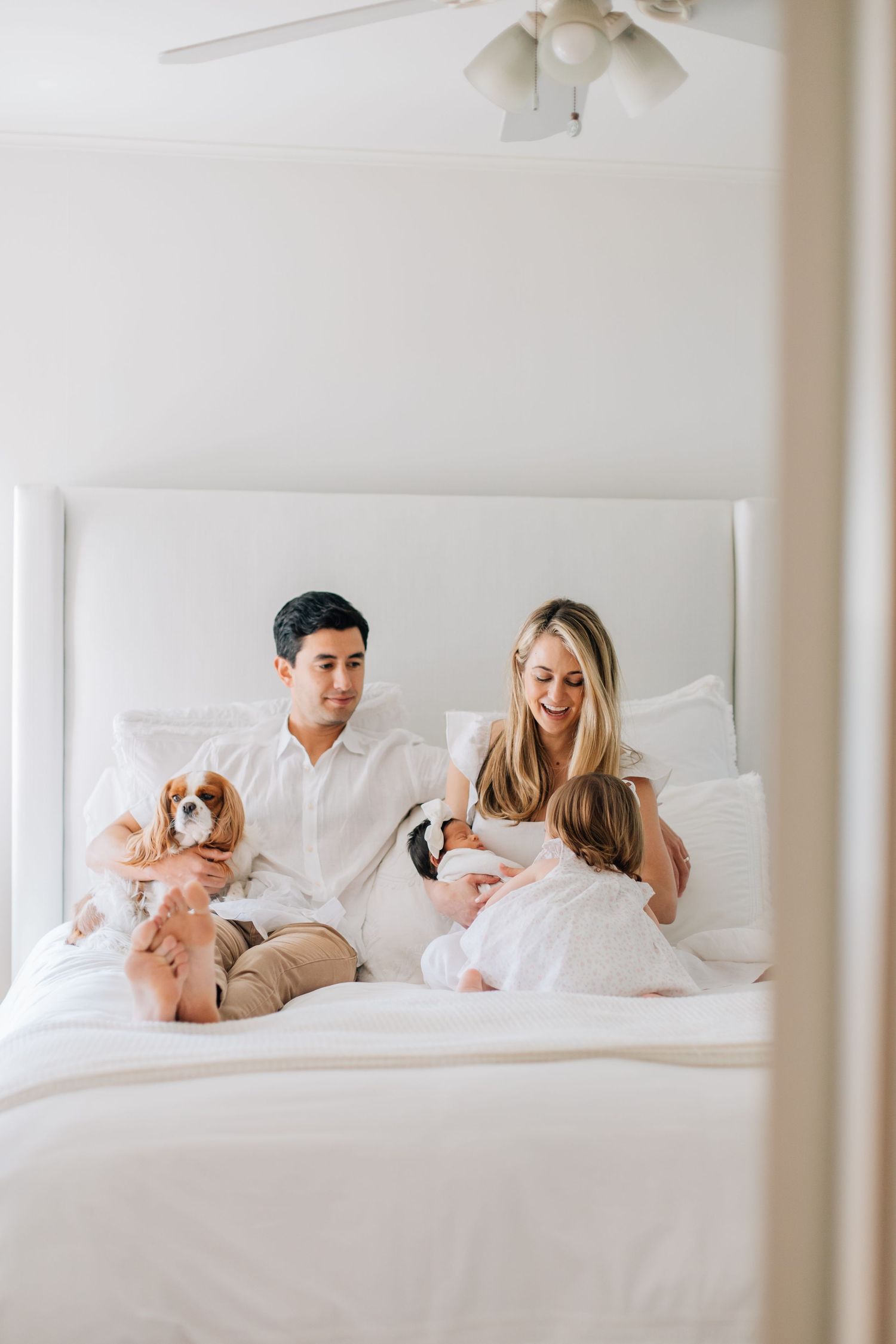 Happy family with father holding dog, mother, and toddler daughter relaxing together on white bed.