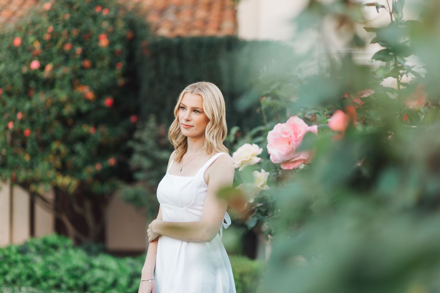 Blonde woman in white dress stands pensively beside blooming pink roses in lush garden setting.