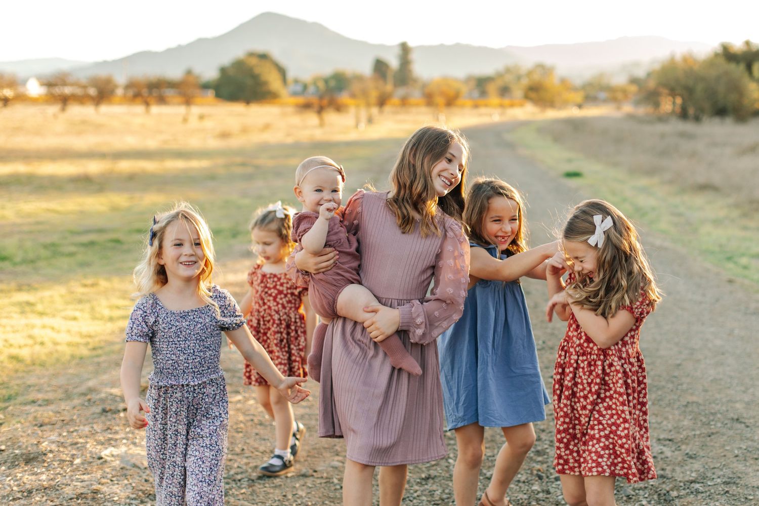 A mother walks joyfully with six young daughters in colorful dresses across a sunlit open field.