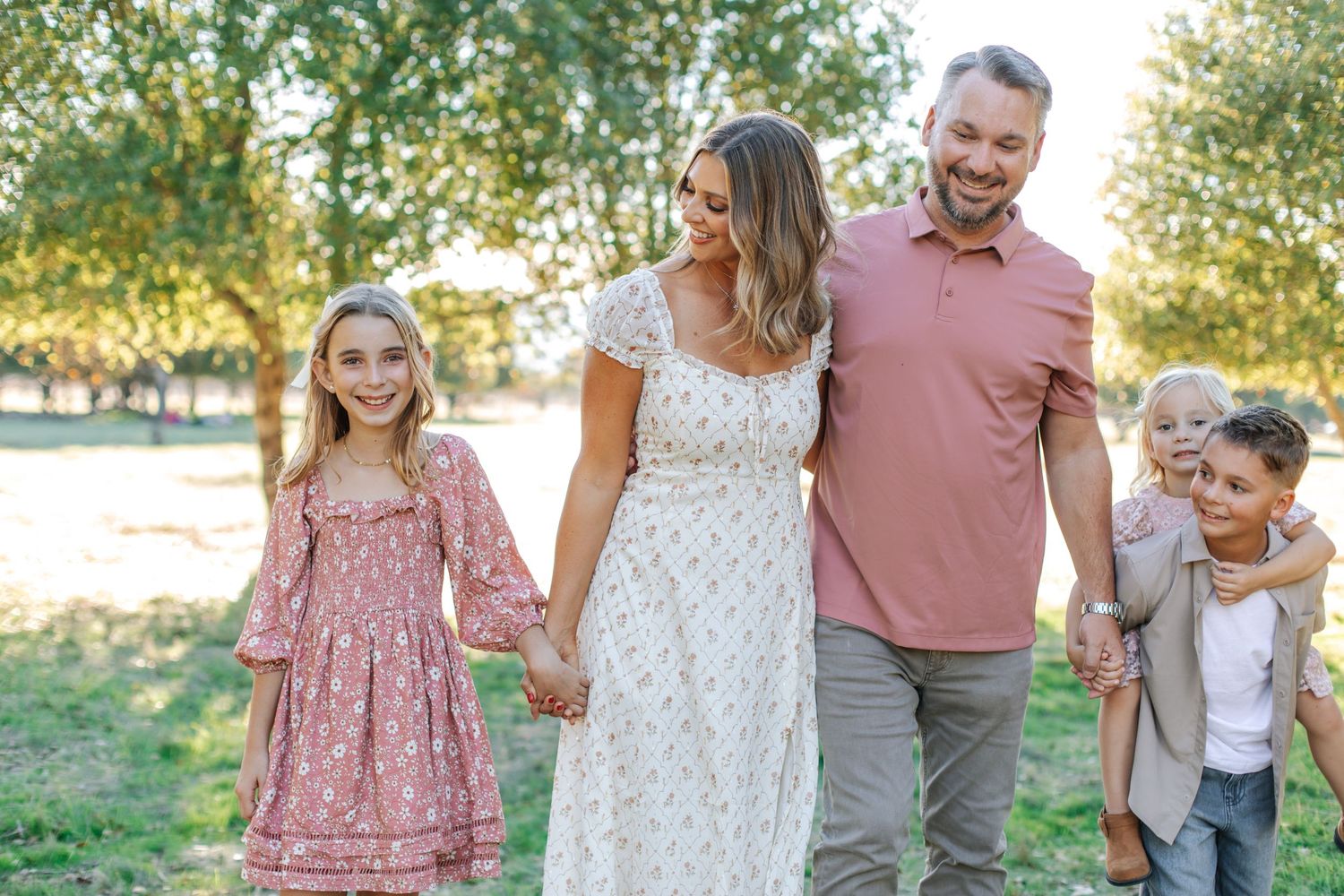 Happy family of four walking outdoors in a sunny park, parents holding hands with two young children.