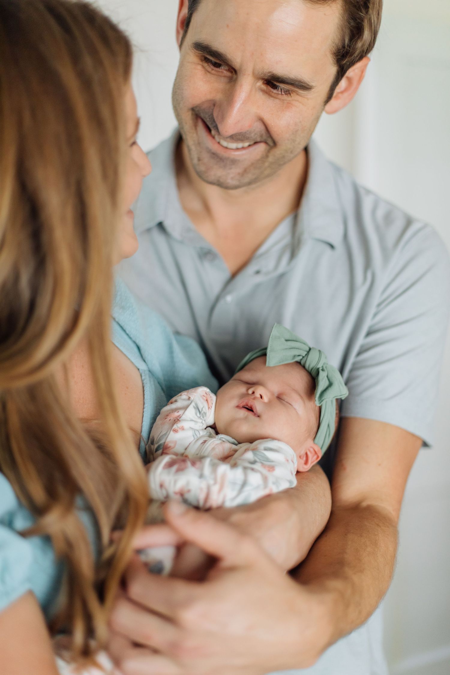Smiling father and mother lovingly gazing at sleeping newborn baby with green bow headband.