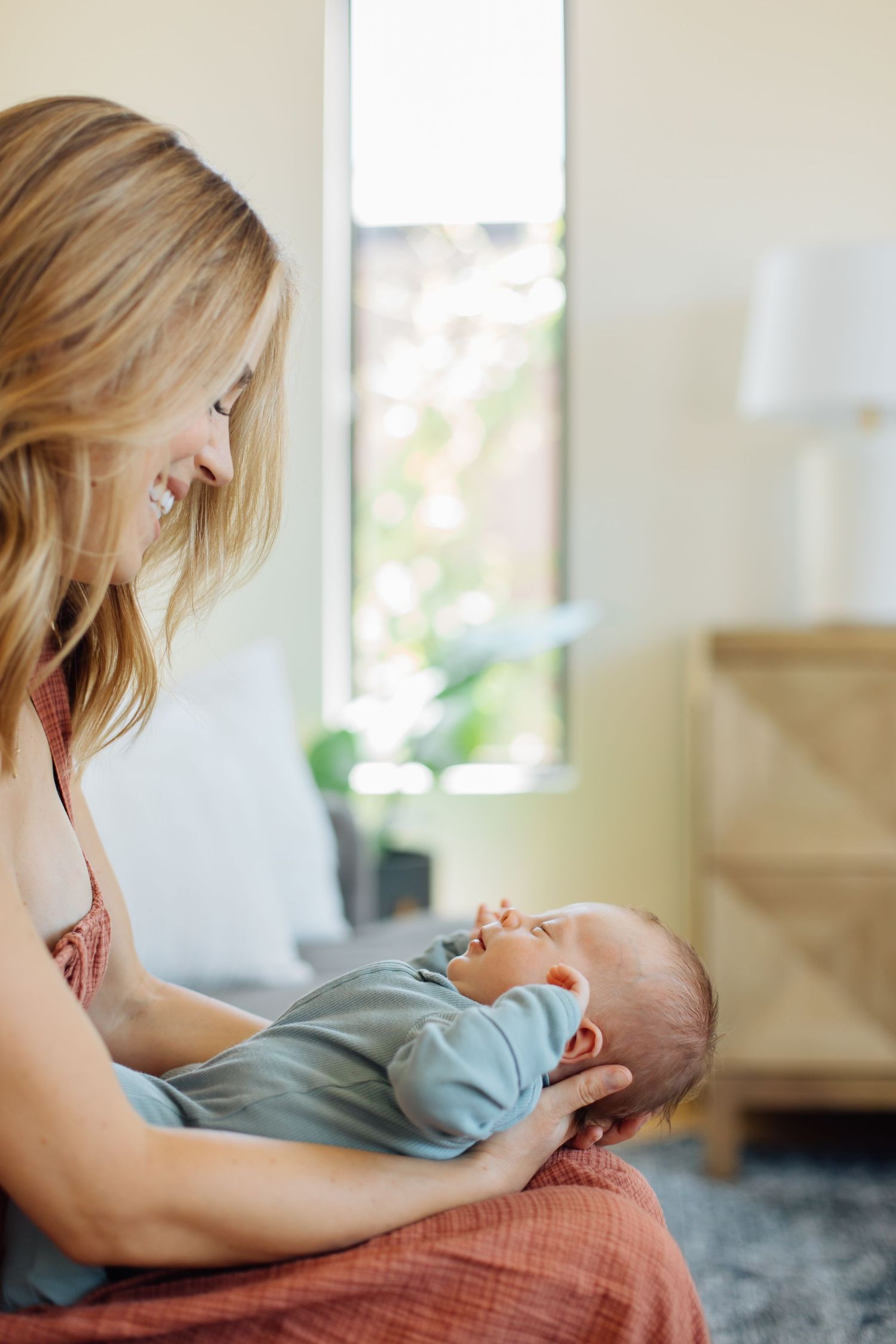 Joyful mother smiling at her newborn baby lying in her arms near a sunny window.