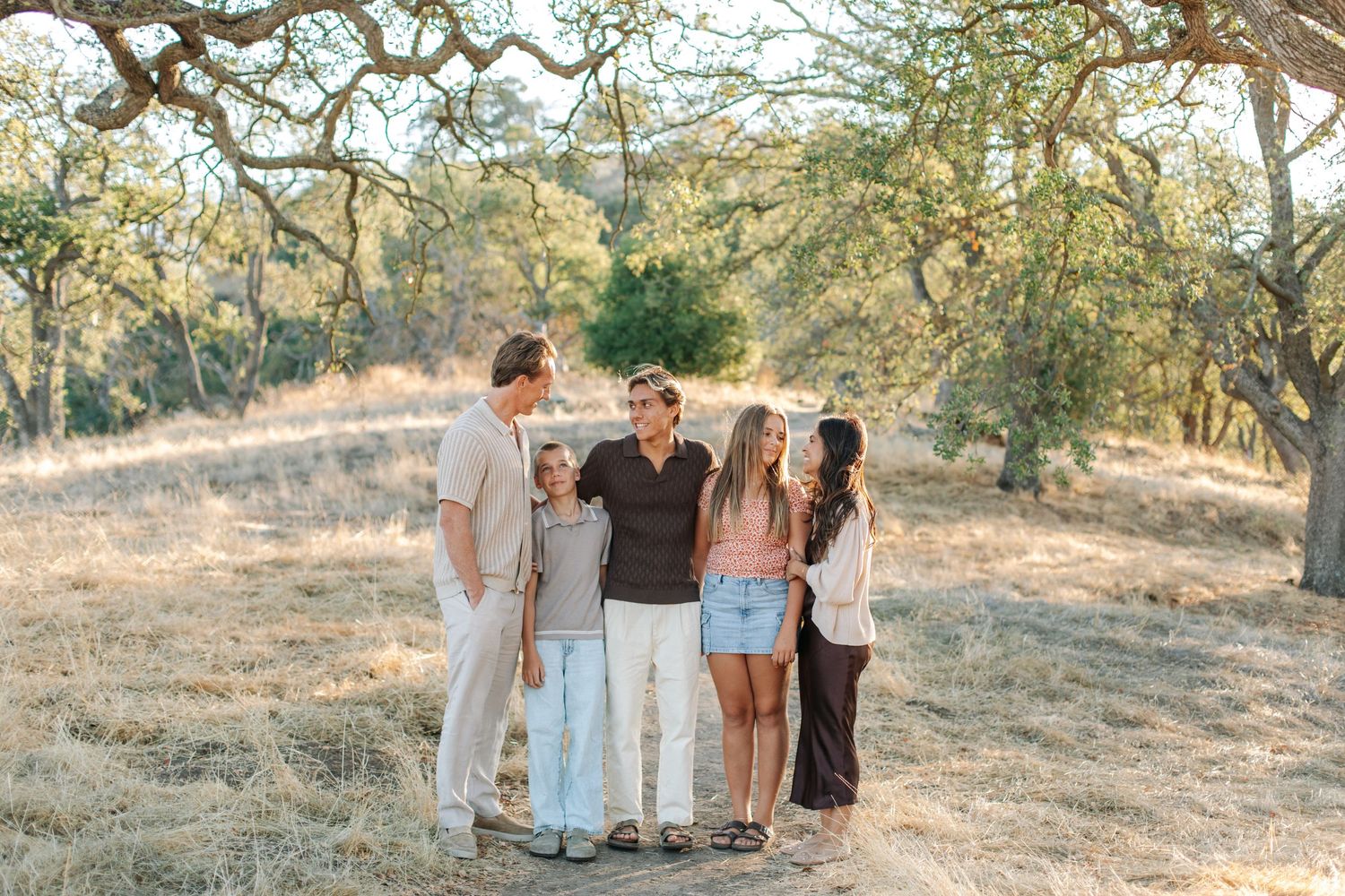 Group of five adults standing together casually on a dry sunlit trail surrounded by oak trees.