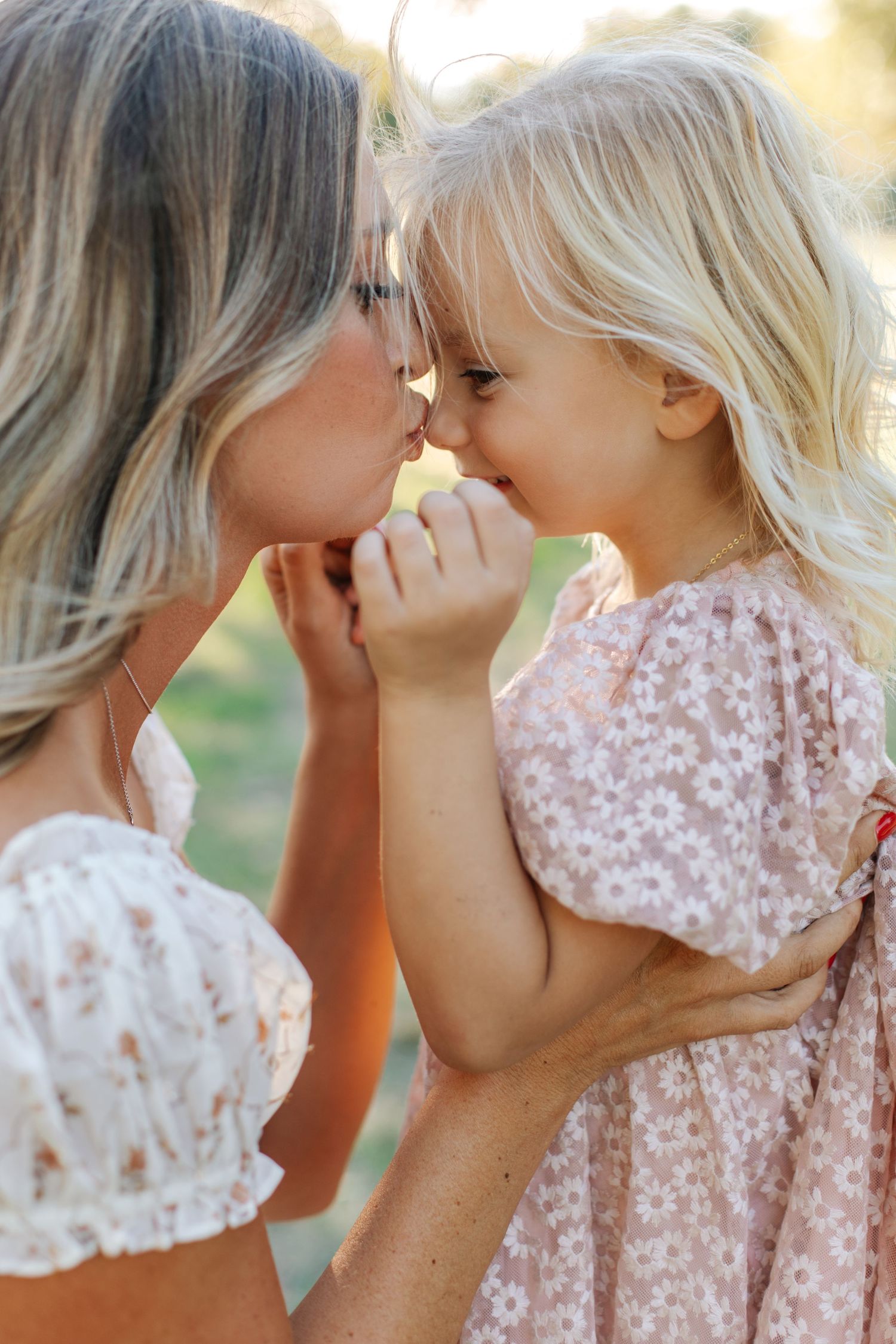 Mother and young blonde daughter touching noses and smiling in a sweet outdoor moment.