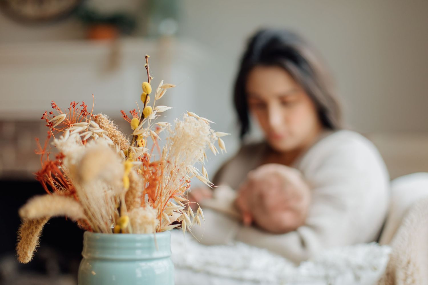 Mother breastfeeding newborn baby on couch with dried flower arrangement in foreground.