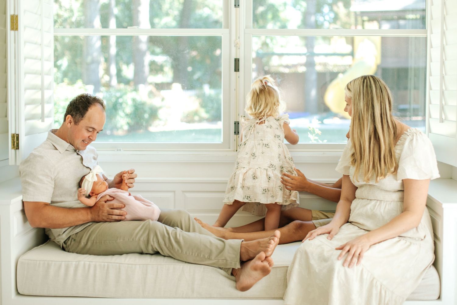 Family relaxes on a white sofa, father holding newborn while toddler plays with mother nearby.