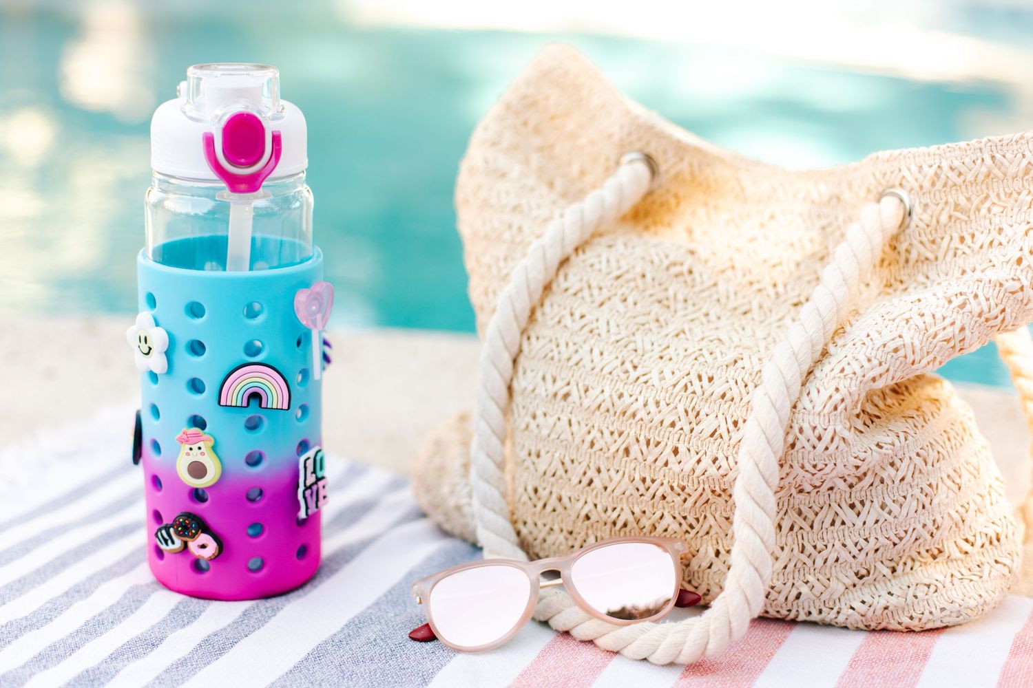 Blue and pink ombre charm water bottle with rainbow stickers beside straw hat near a pool.