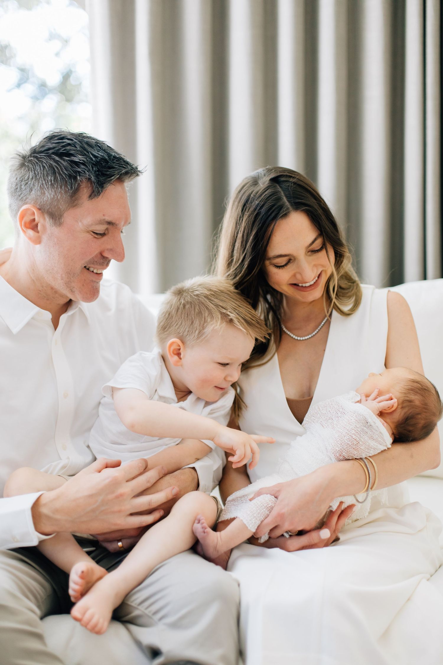 Happy family with father, mother, toddler son, and newborn baby sitting together on a white sofa.