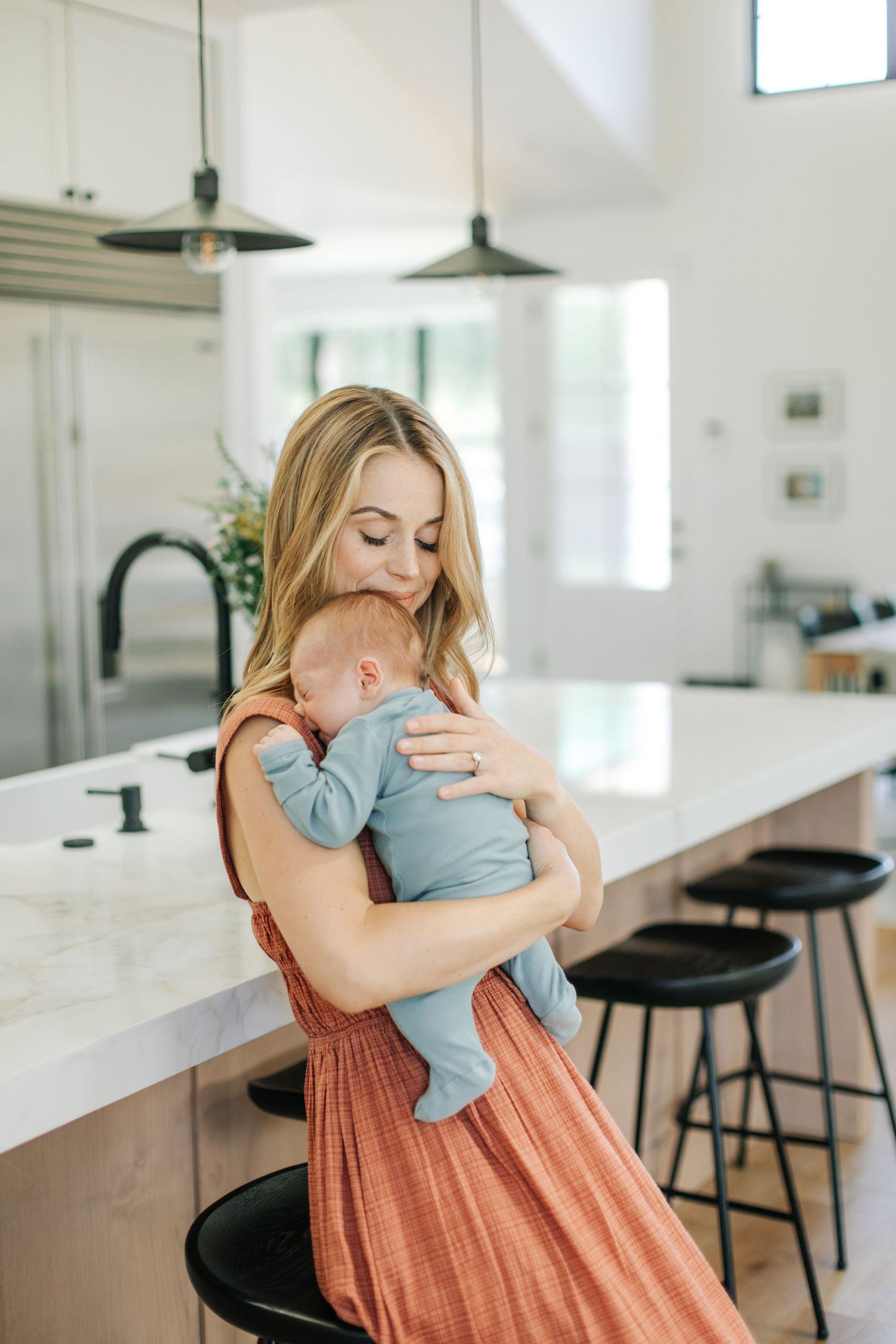 Blonde mother in terracotta dress cradling sleeping newborn baby near white marble kitchen island.
