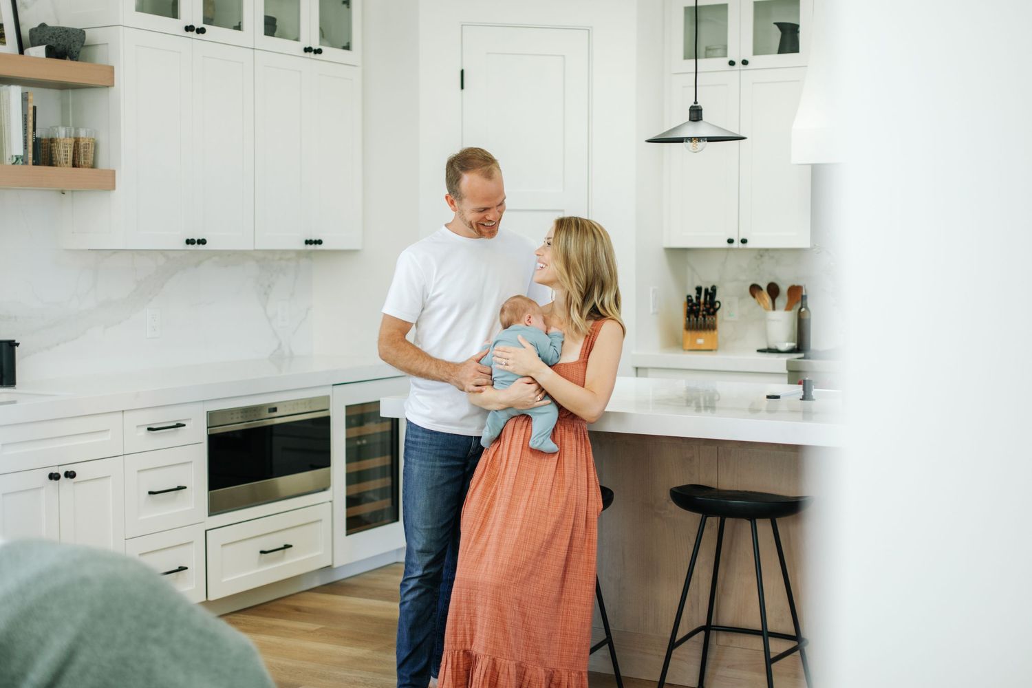 Happy couple holding newborn baby in a bright modern white kitchen at home.