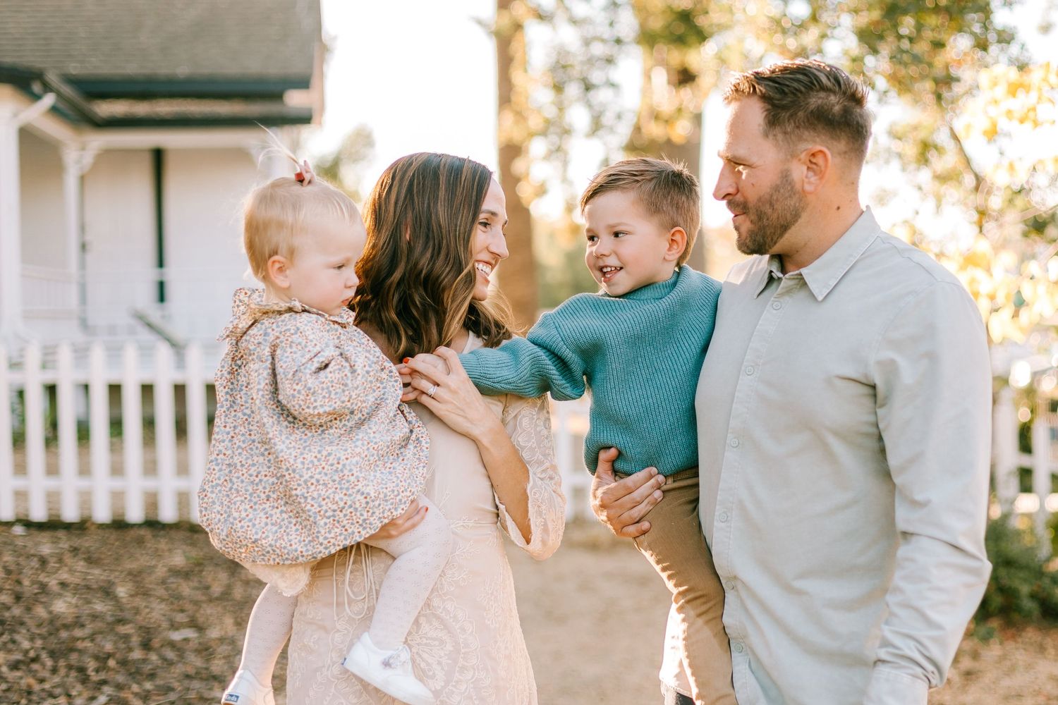 Happy young family with two toddlers smiling outdoors near white picket fence.