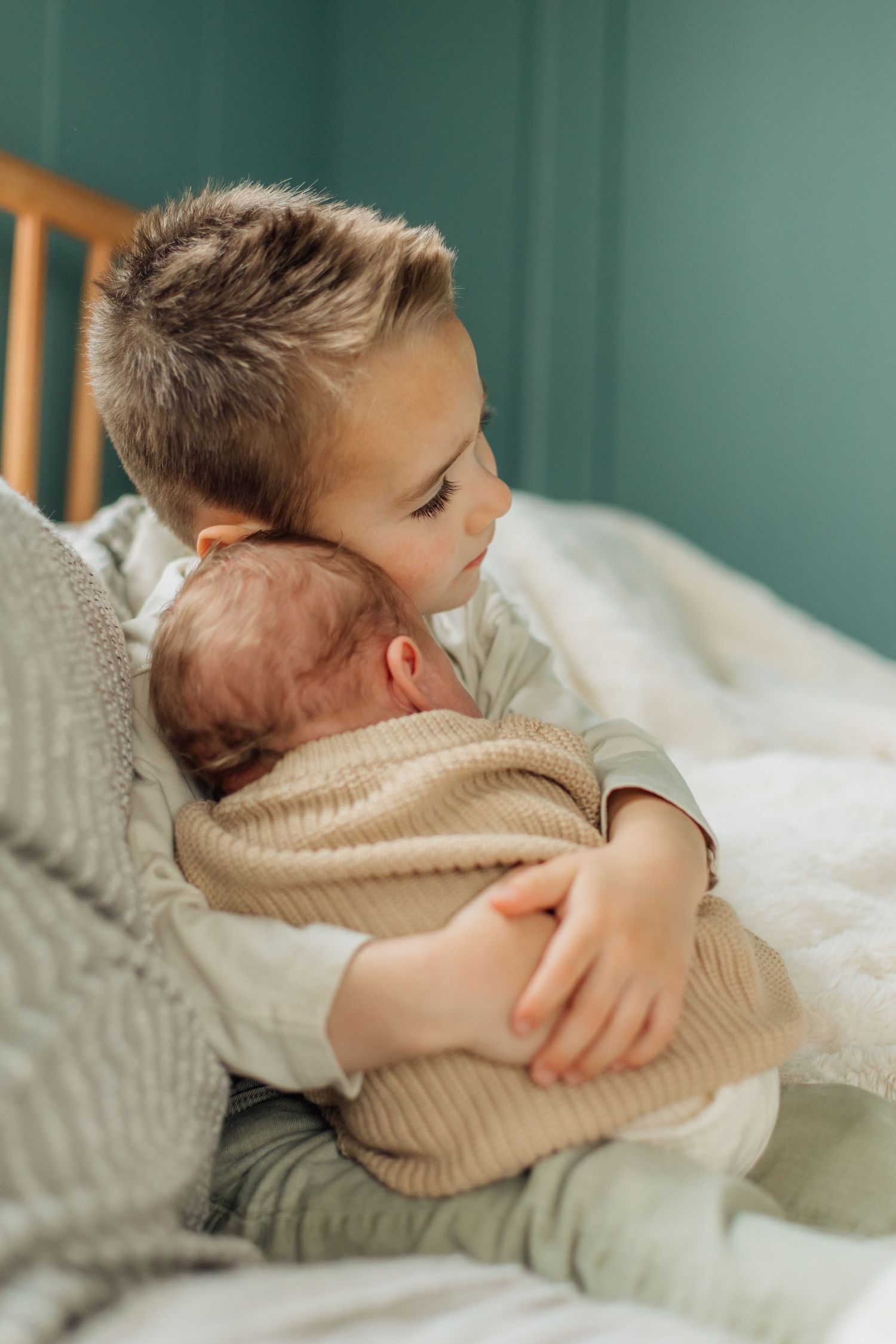 Older brother gently kissing newborn baby while sitting together on white bedding.