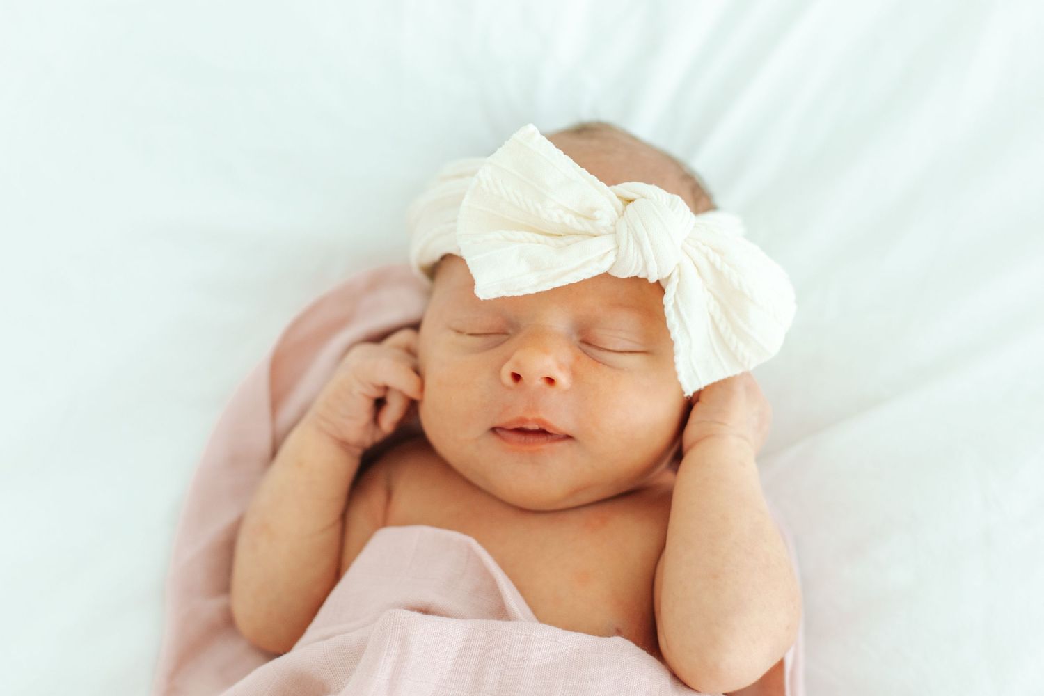 Peaceful newborn baby girl sleeps wearing a white bow headband, wrapped in a soft pink blanket.
