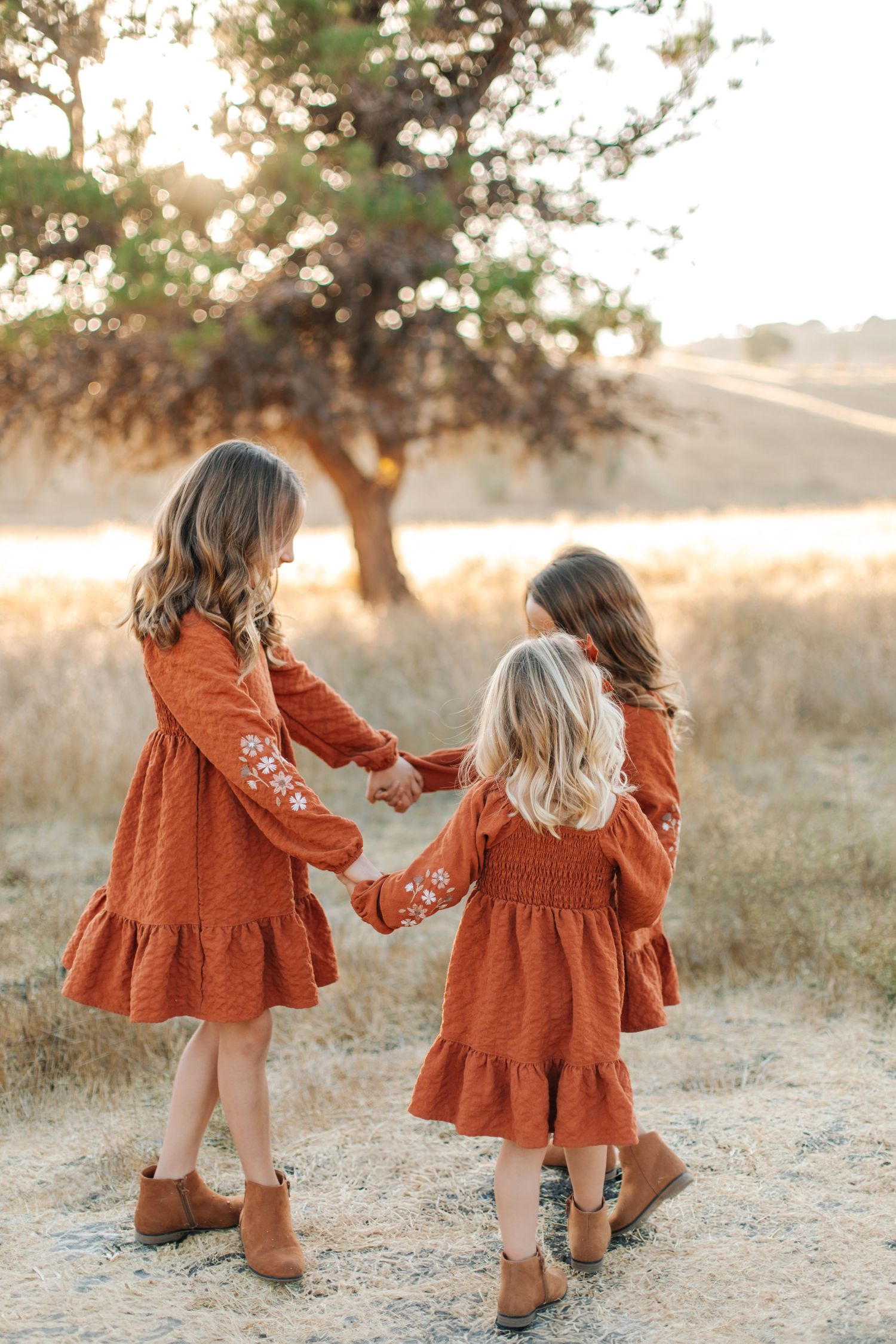 Two young sisters in matching rust dresses hold hands walking through a golden sunlit field.