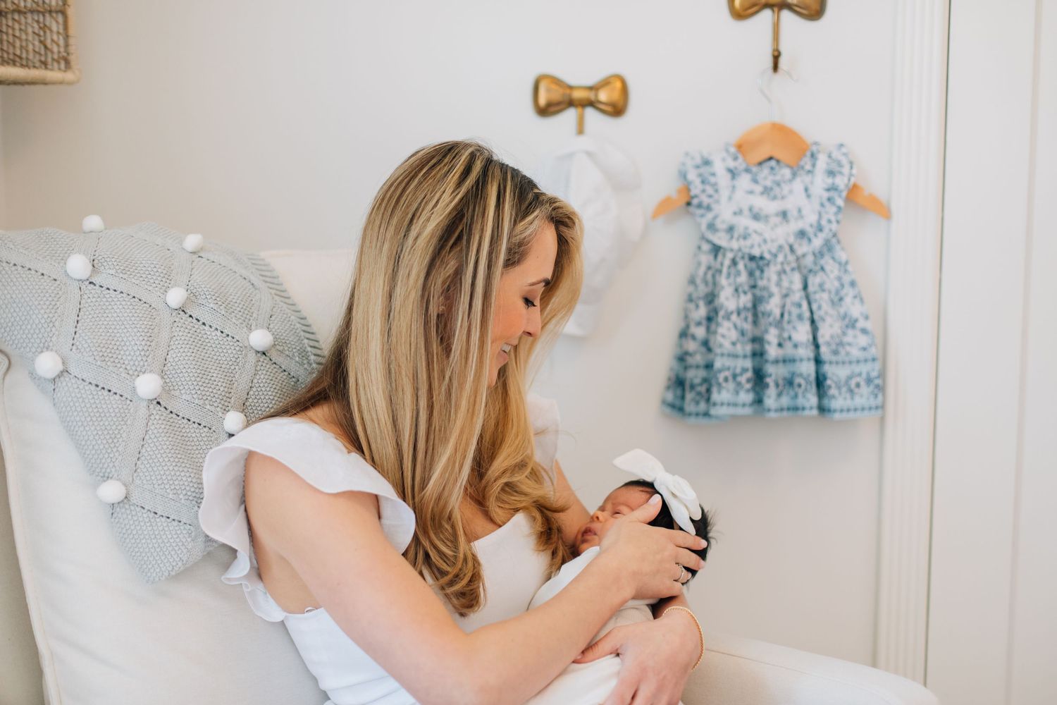 Blonde mother in white dress lovingly gazes at her swaddled newborn in a softly decorated nursery.