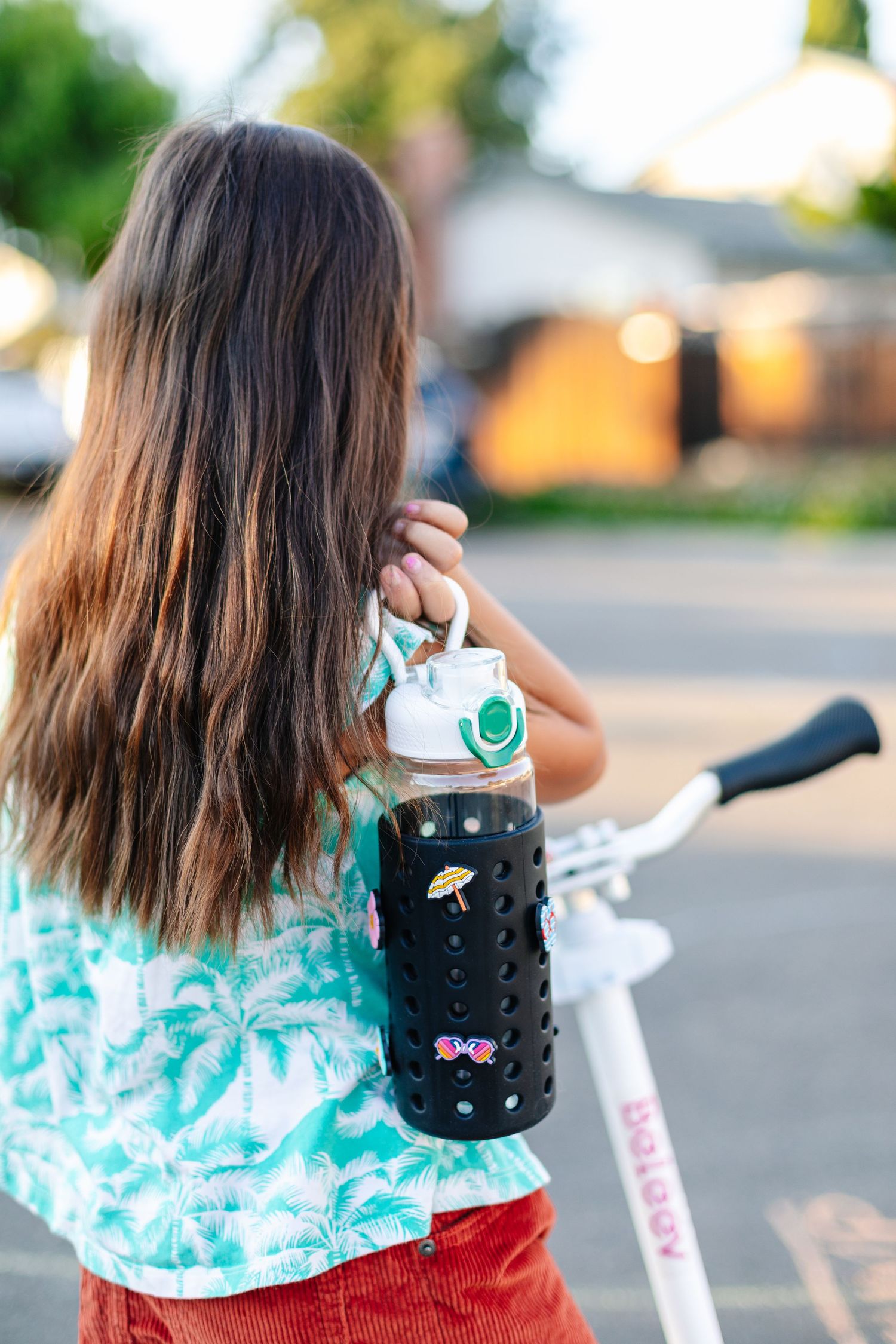 Girl with long brown hair carrying a black silicone-sleeved glass water bottle near a white bike.