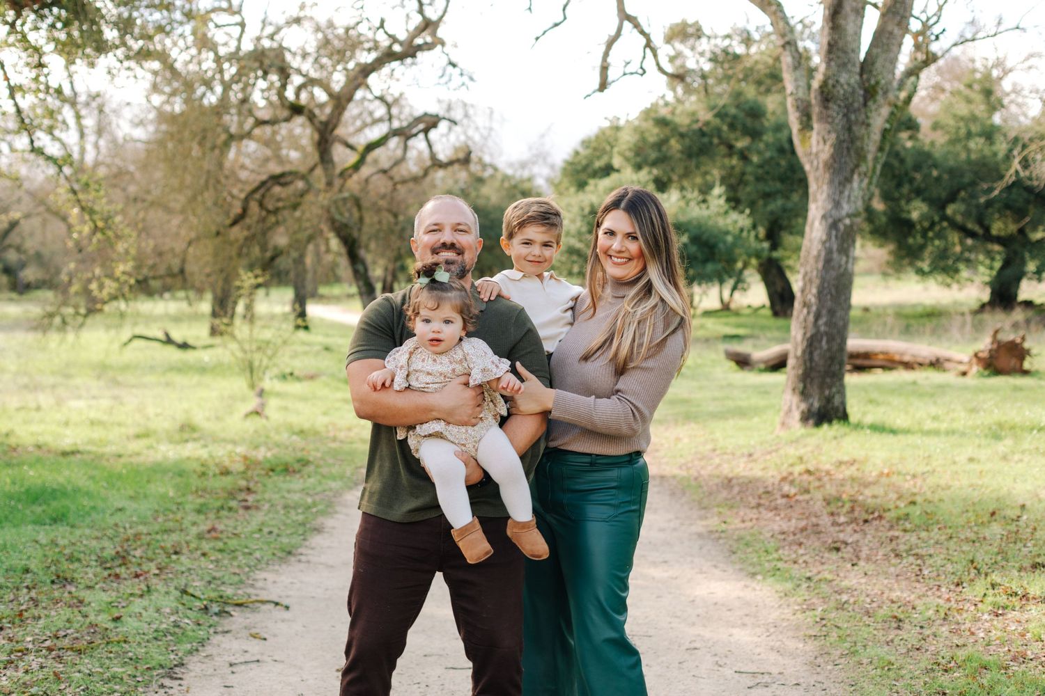 Happy family of four posing outdoors on a scenic park path surrounded by lush green trees.