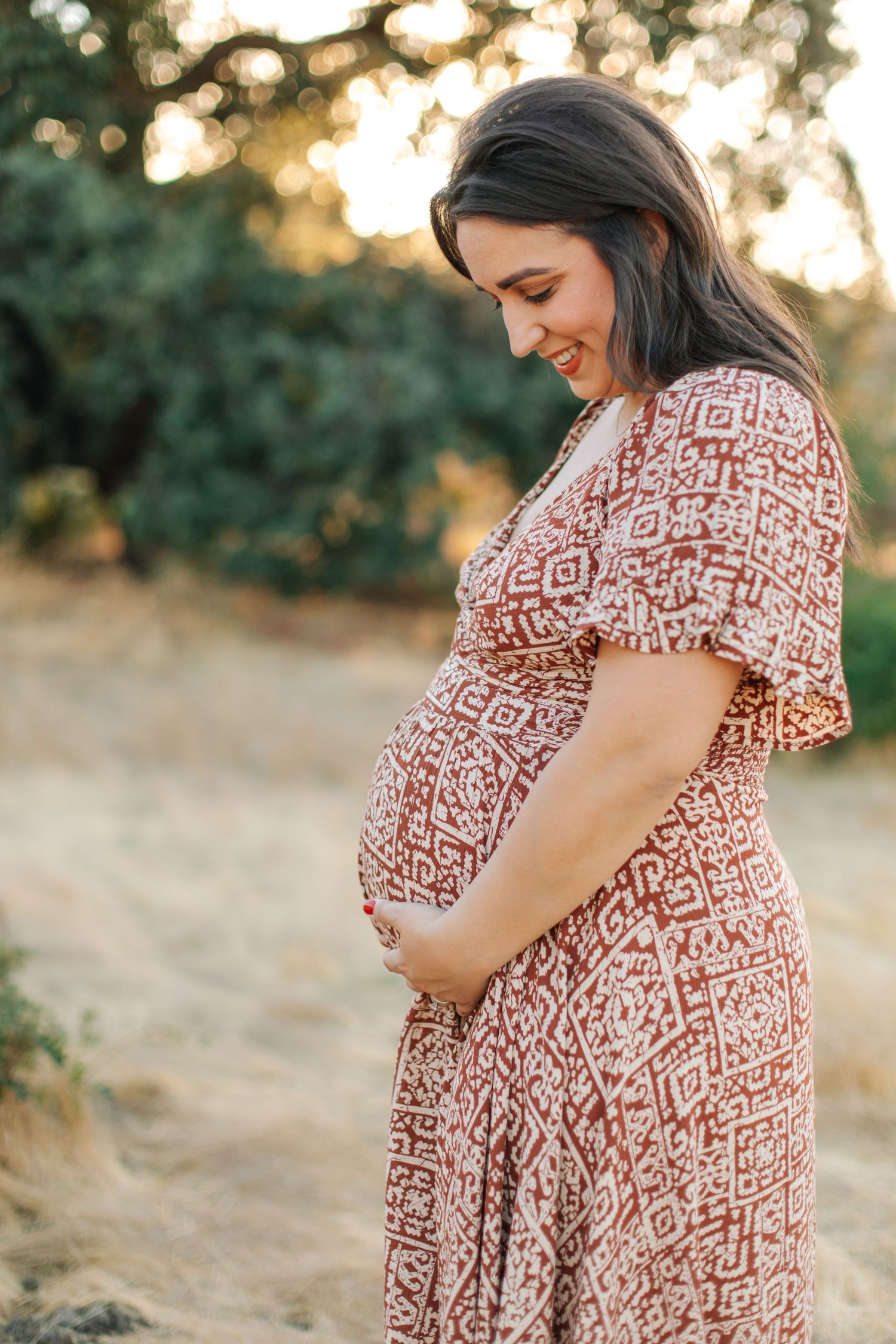 Pregnant woman in boho red patterned dress smiling down at her baby bump in golden light.