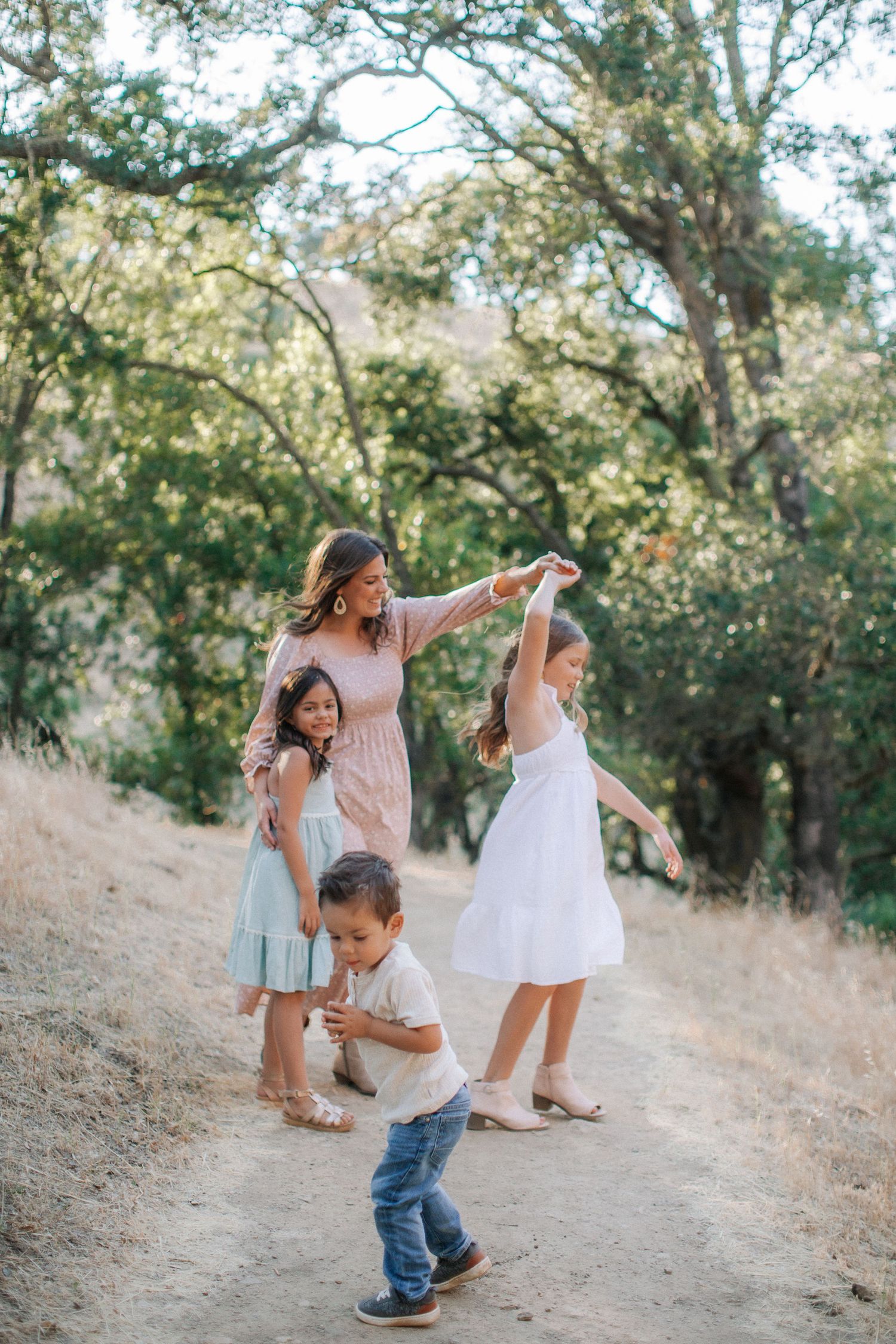 Mother playfully dancing and spinning with her three young children on a shaded outdoor dirt path.