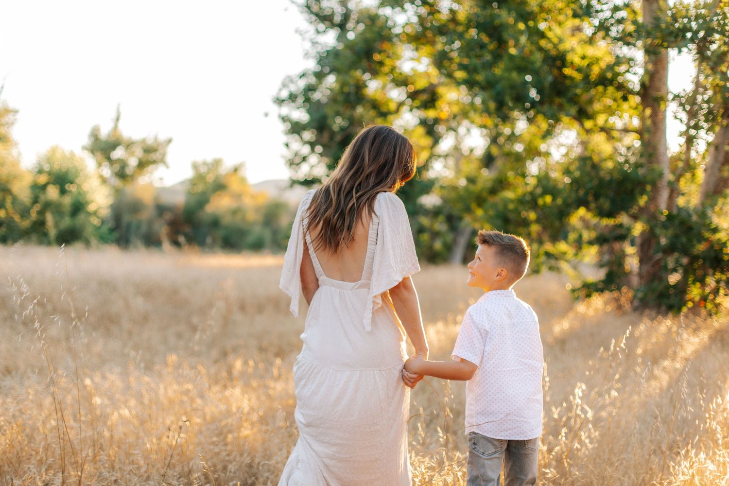 A woman in a white dress holds hands with a young boy while walking through a golden sunlit field.
