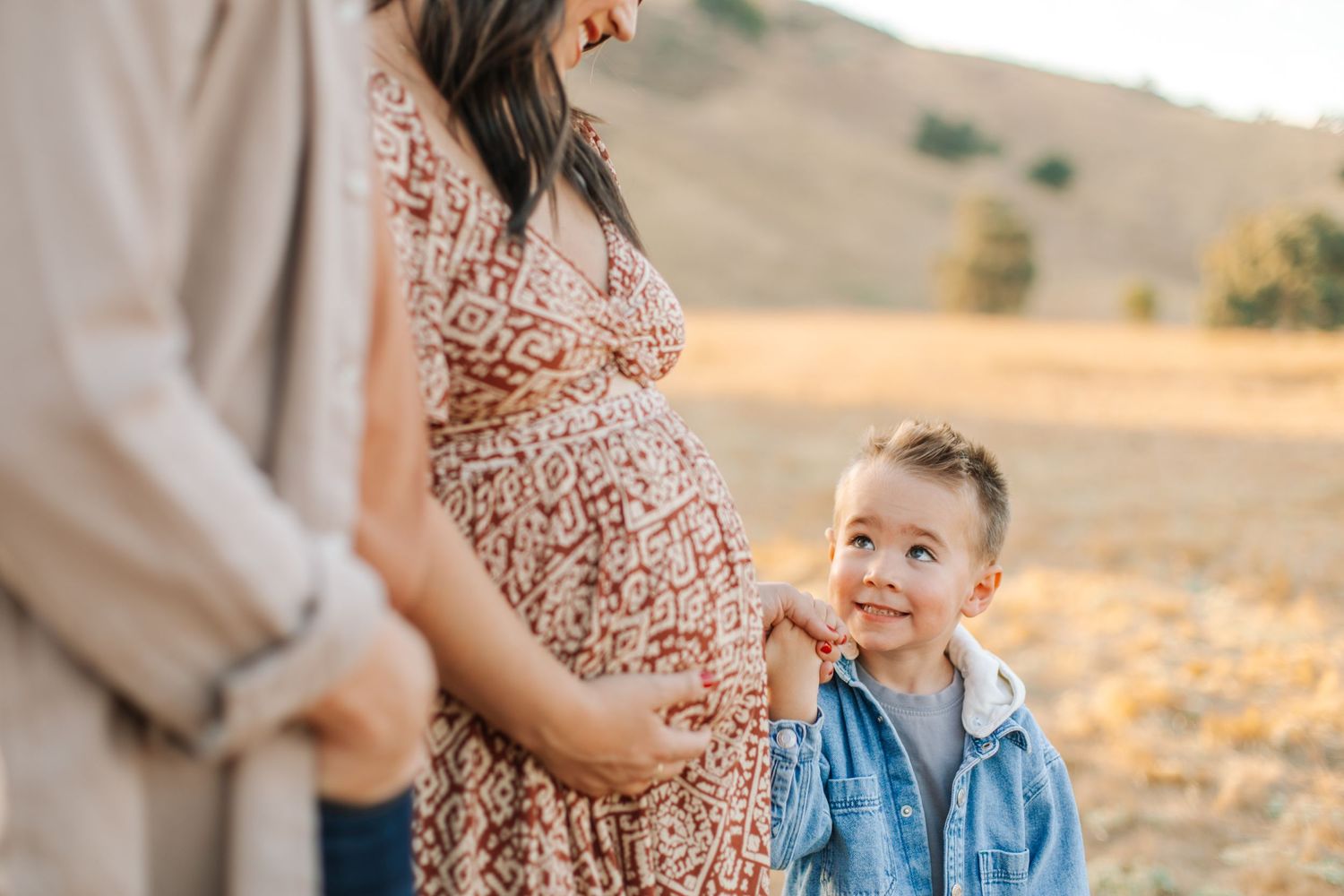 Young boy smiling up at pregnant mother while touching her baby bump in a sunny field.