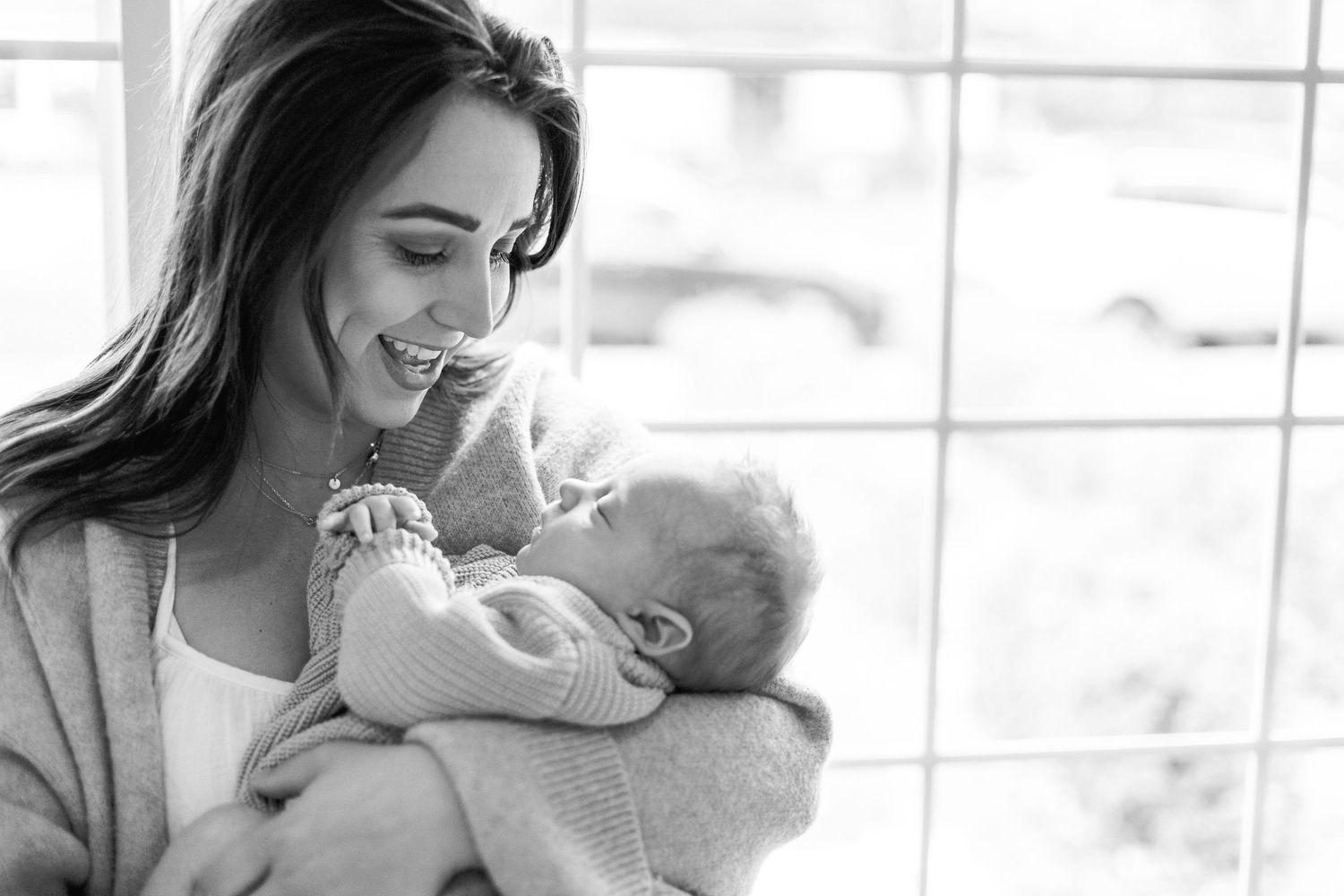 A black and white portrait of a smiling mother tenderly holding her sleeping newborn by a window.