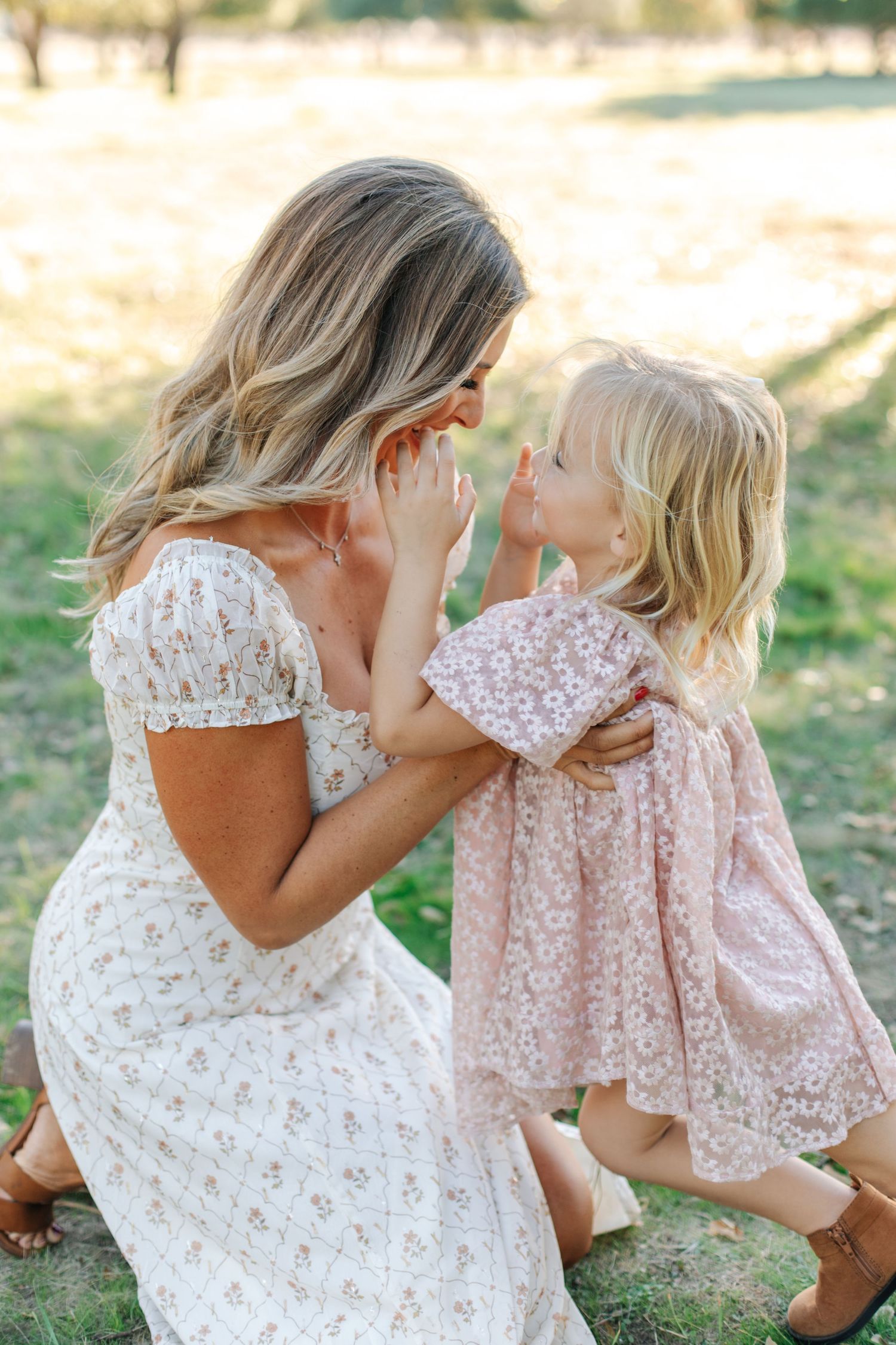 Blonde mother in white floral dress crouching to interact with her young daughter in pink dress.