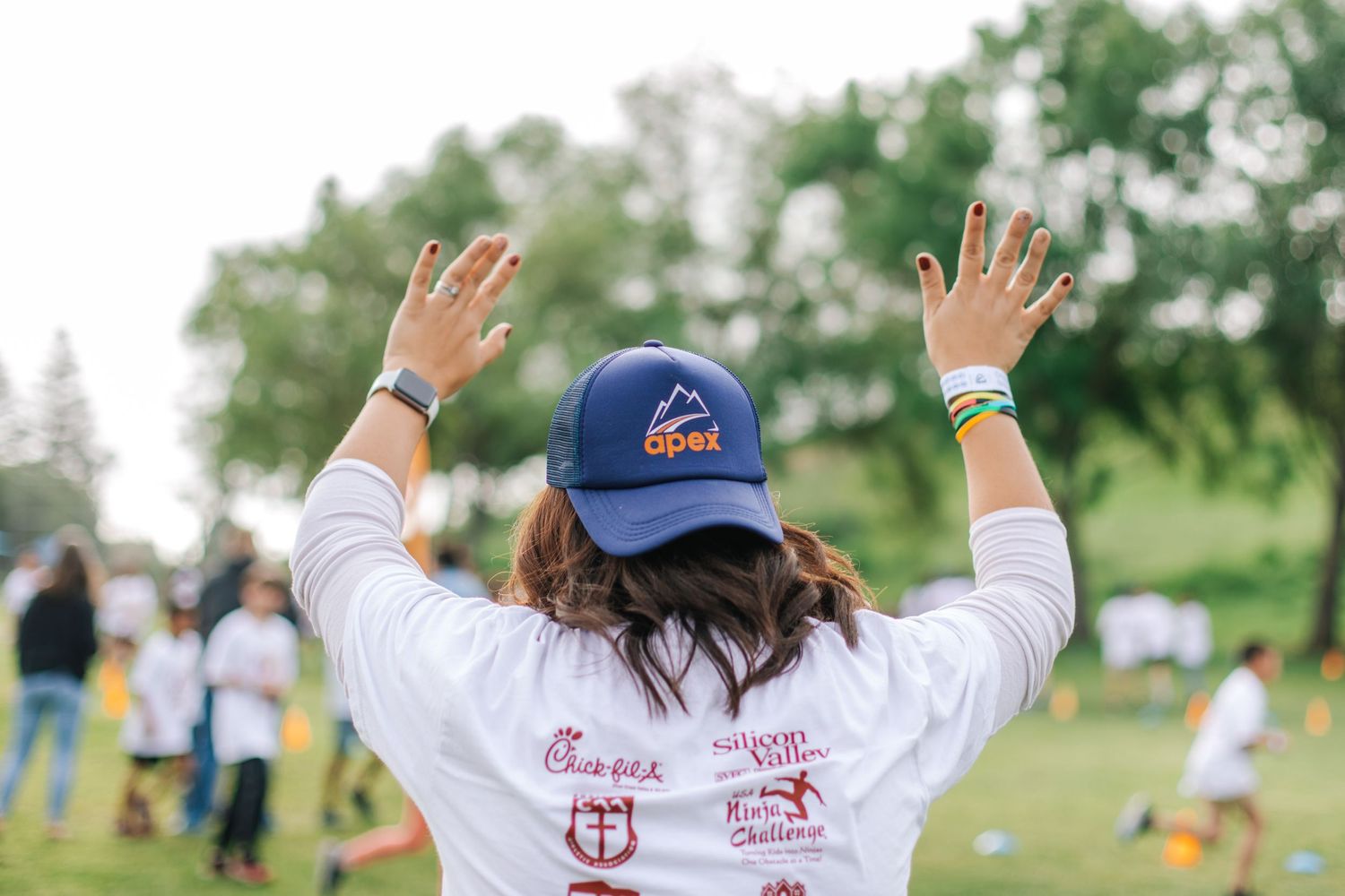 Woman in Apex cap and white shirt raises hands at outdoor fun run charity event.