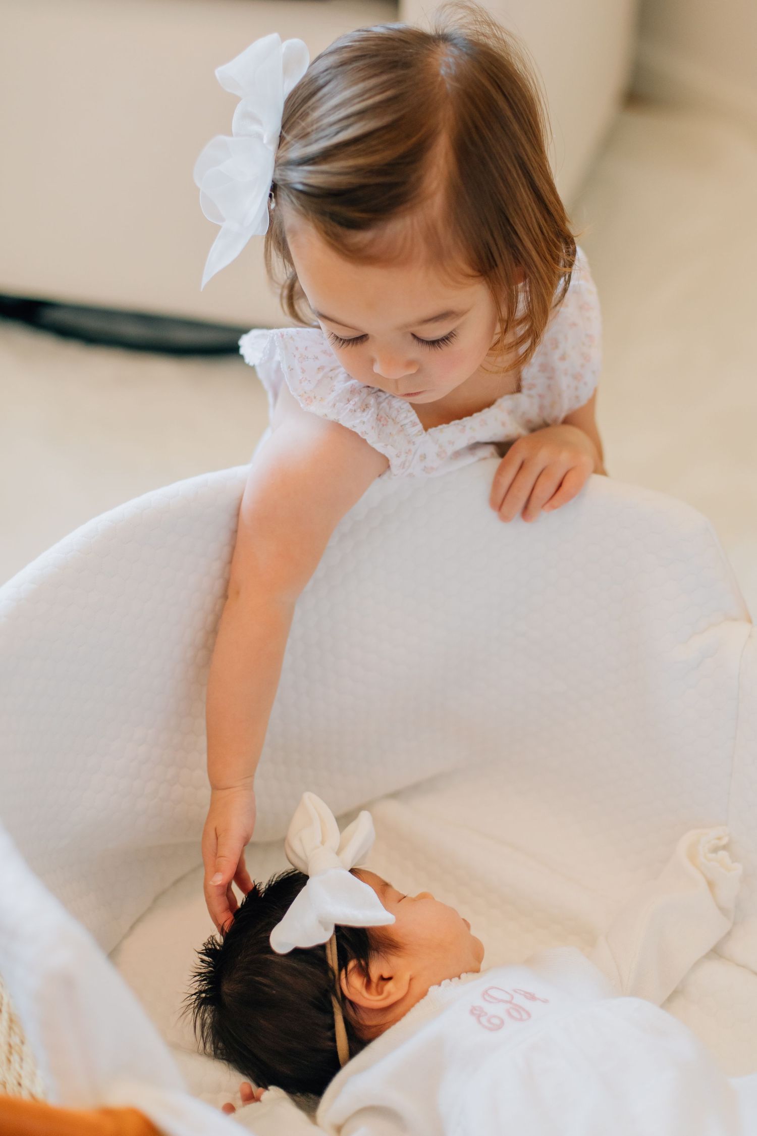 Curious toddler girl with white bow gently touches newborn sibling resting in a white bassinet.