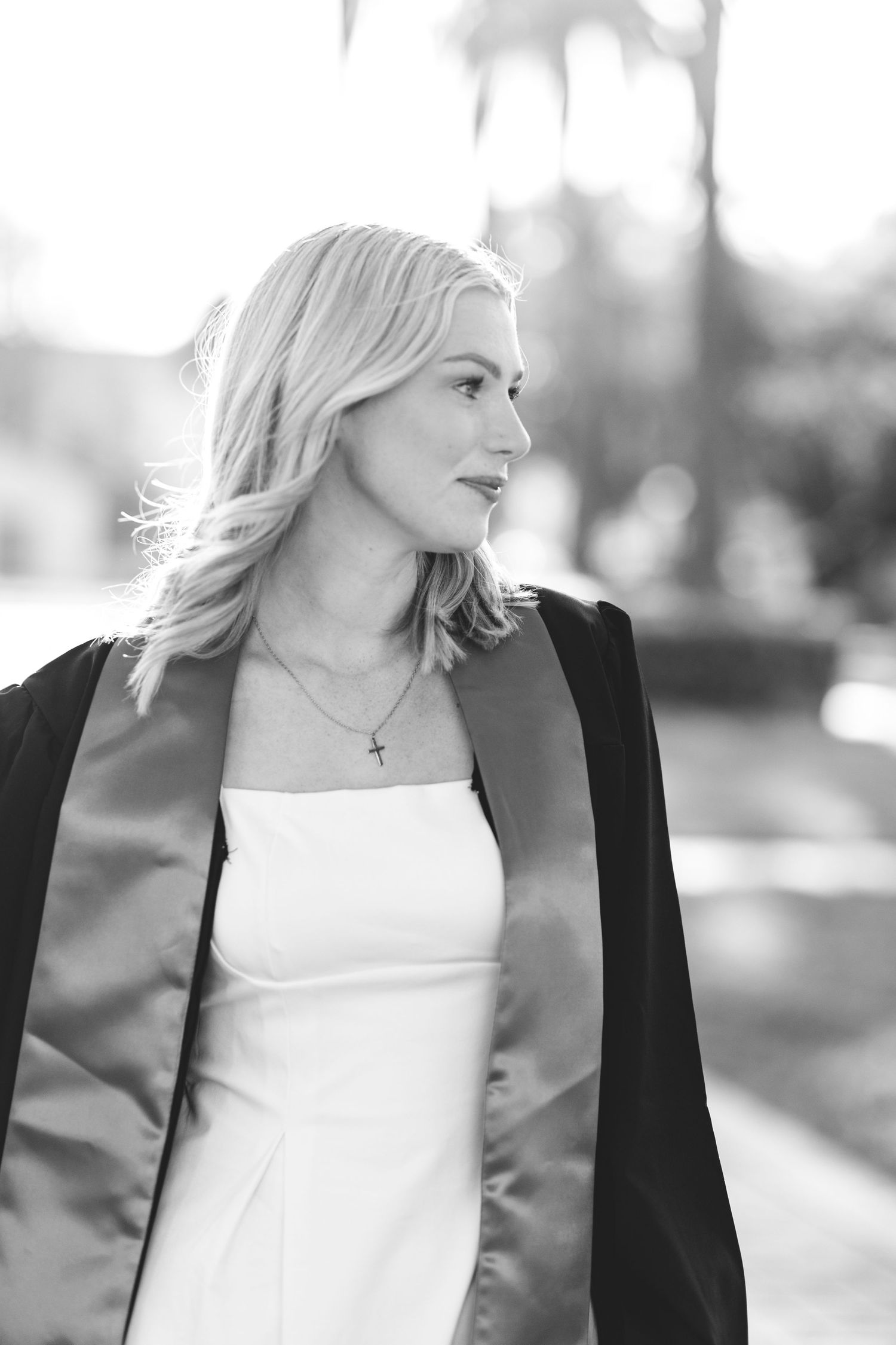 Black and white portrait of blonde woman in graduation gown with cross necklace looking to the side.