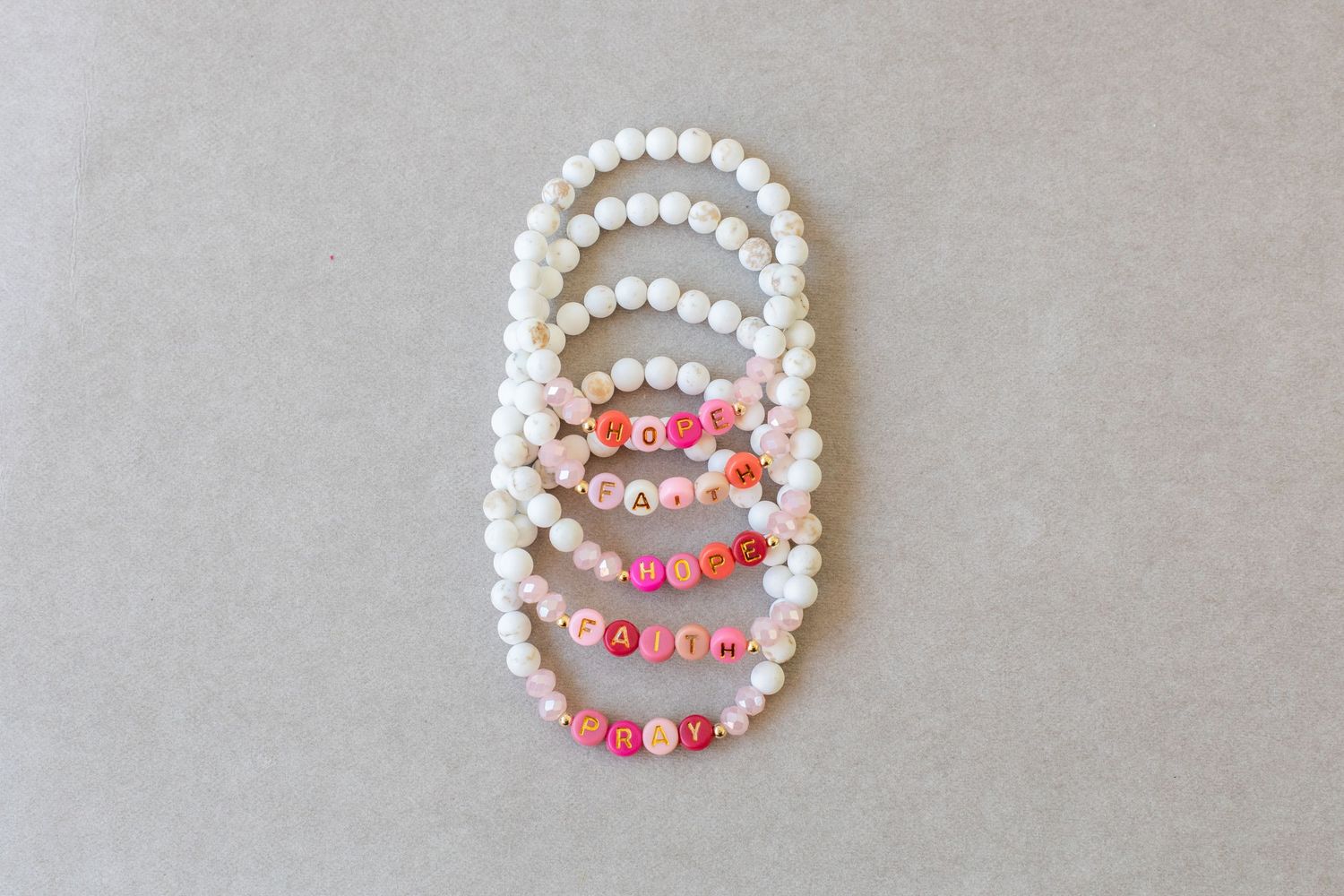 Handmade beaded bracelets with colorful letter beads spelling words on white pearl beads, arranged on gray background.