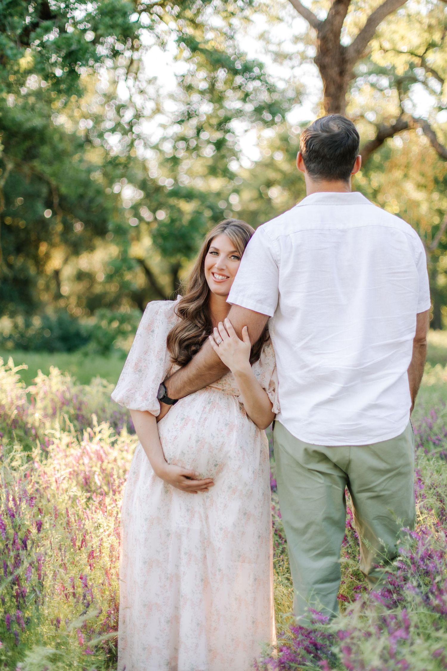 Pregnant woman in floral dress smiling while leaning against partner in white shirt in wildflower field.