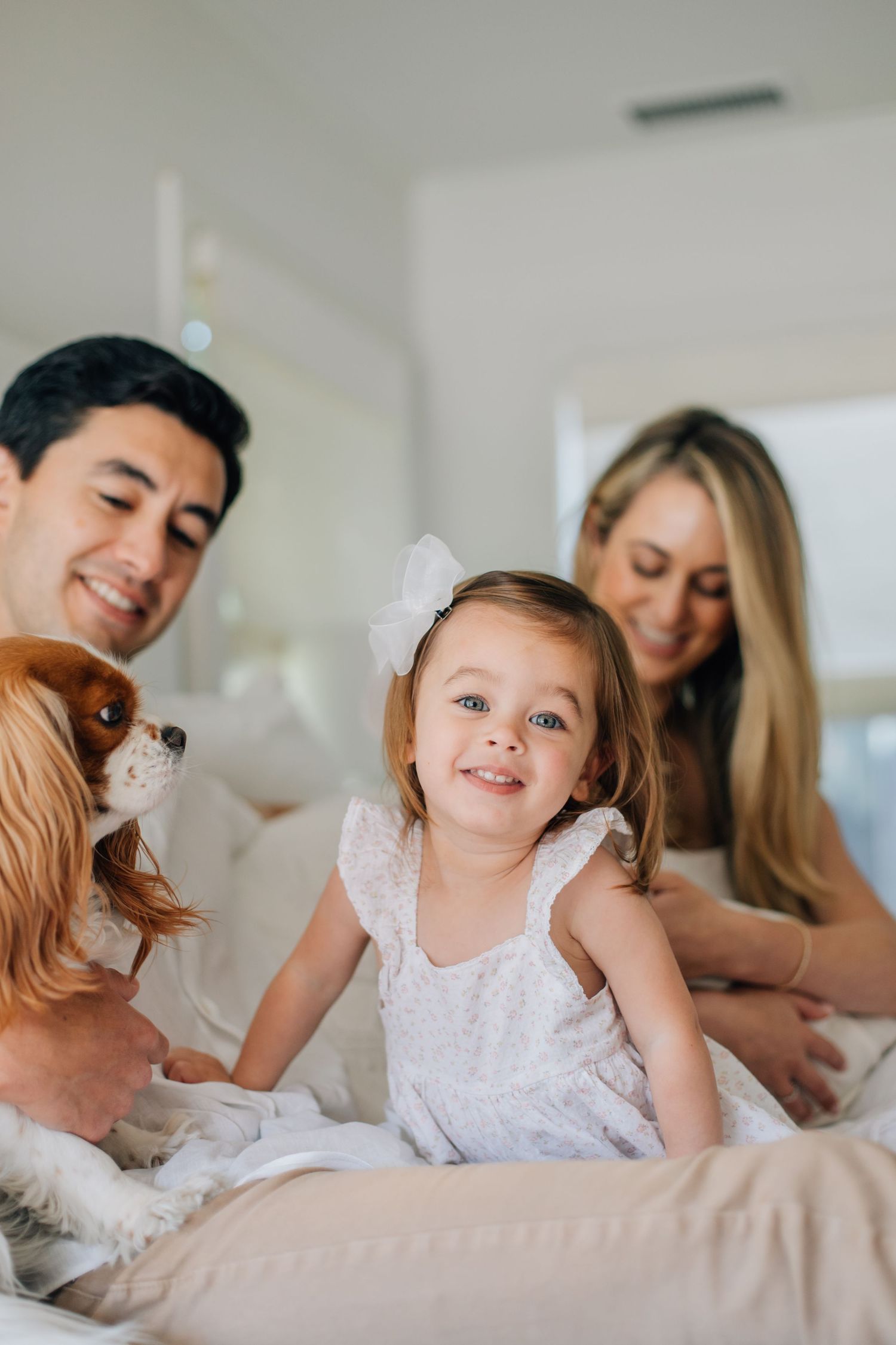 Smiling toddler girl with white bow poses with parents and a Cavalier King Charles Spaniel on bed.