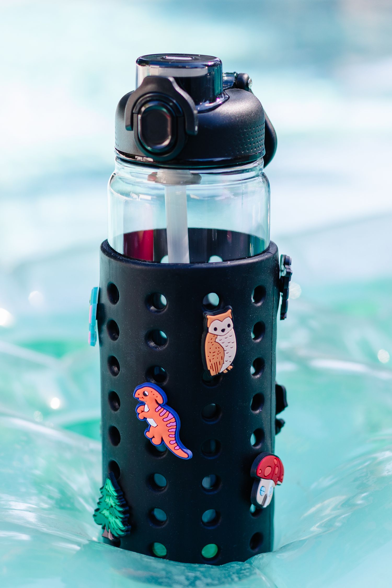 Black silicone sleeve water bottle decorated with owl, dinosaur, and nature charms near a pool.