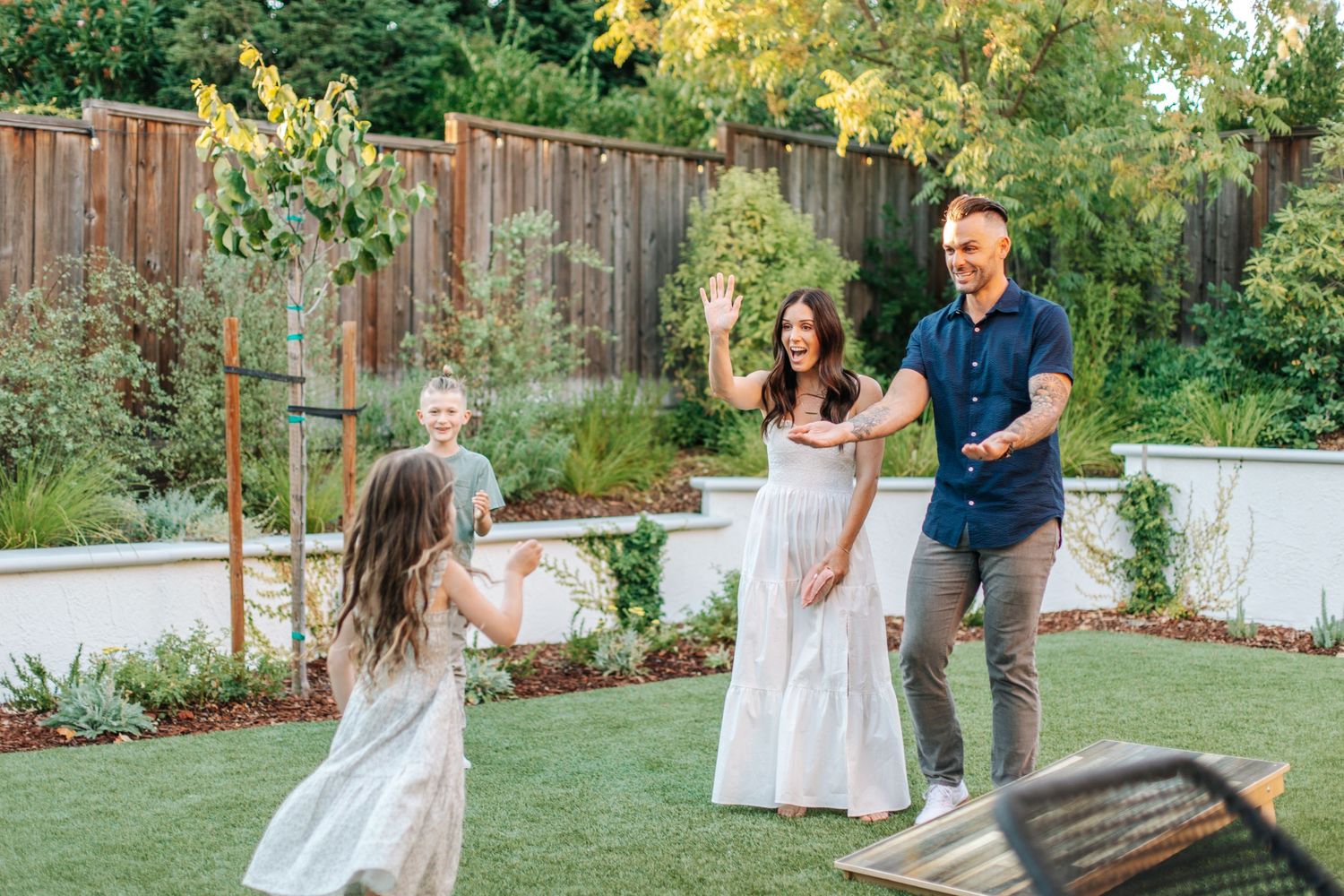 A couple laughs and waves as their daughter runs toward them during a backyard game.