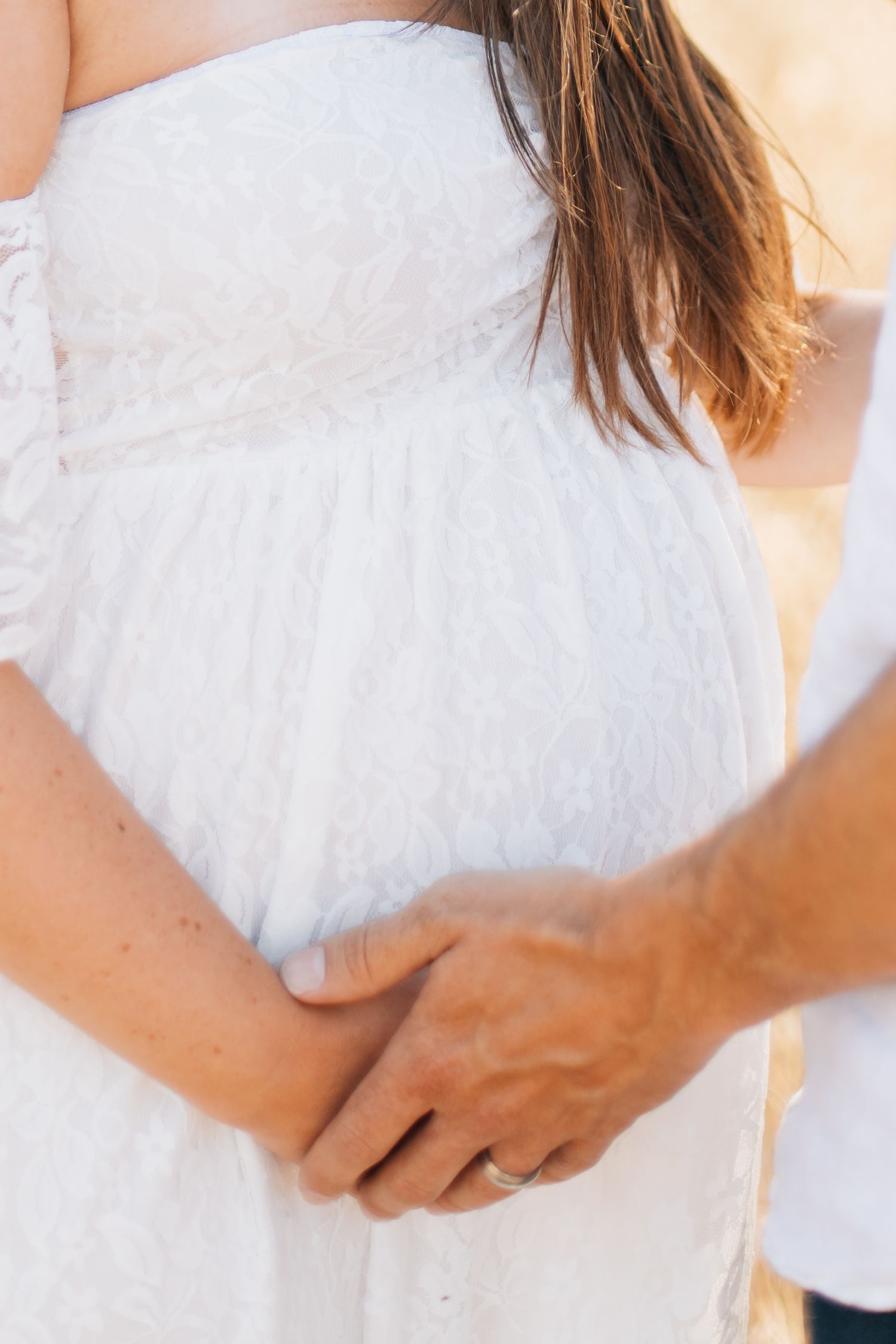 Close-up of pregnant woman in white lace dress with partner's hands gently holding baby bump.