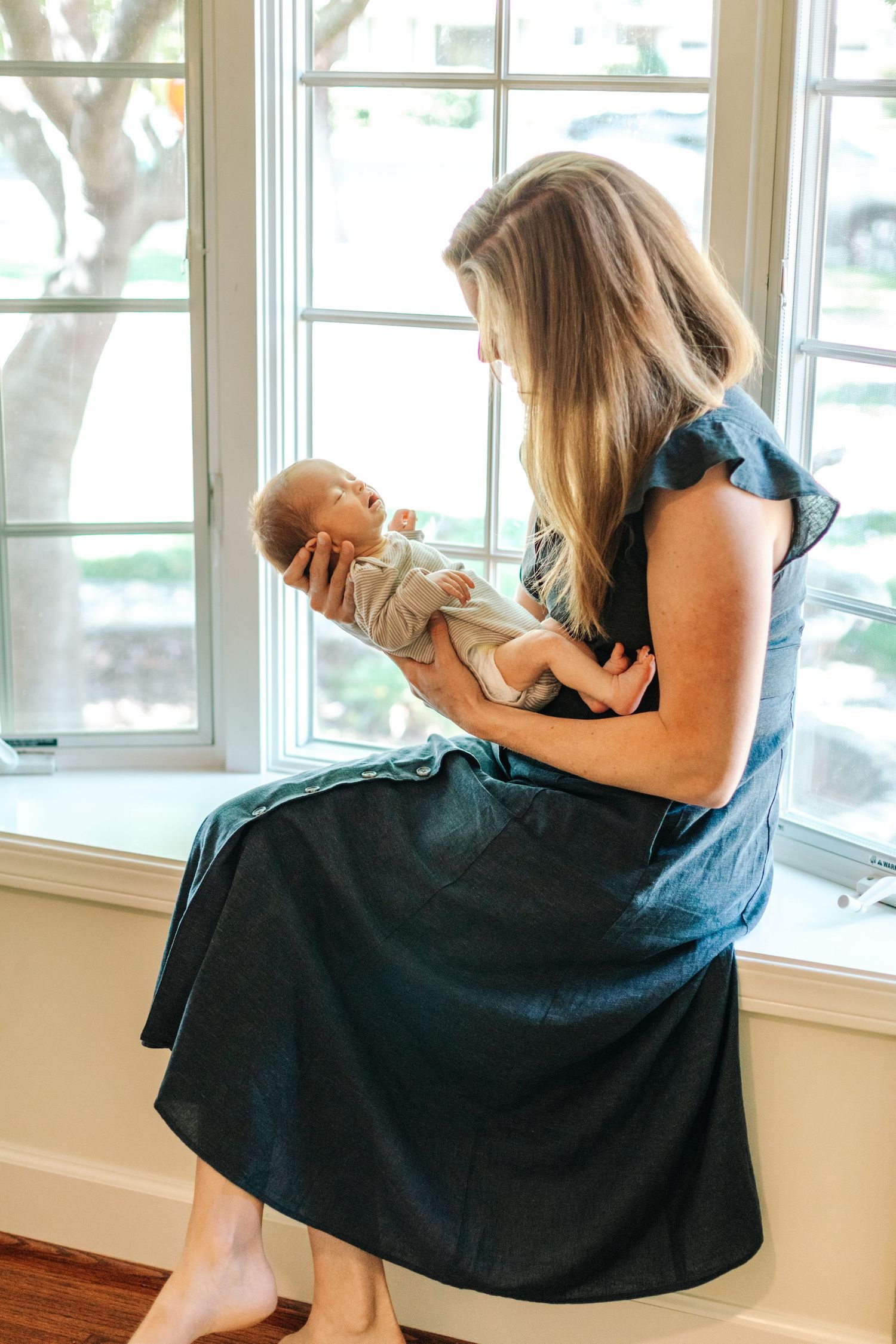 Mother in teal dress sitting by window holding and gazing at her newborn baby in natural light.