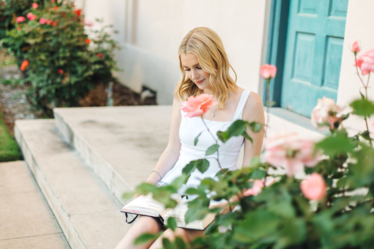 Blonde woman in white dress sitting on steps smelling a pink rose beside a teal door.