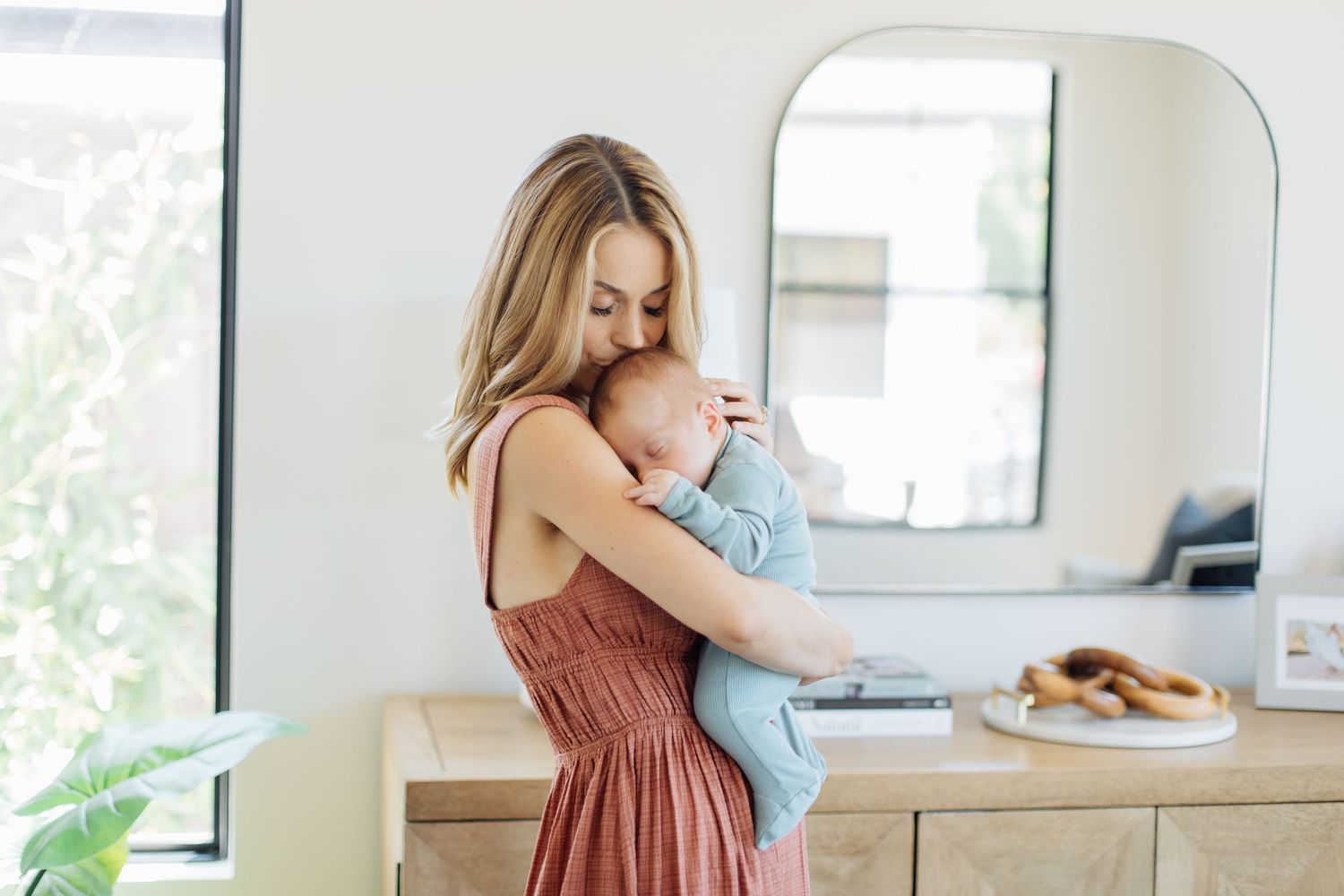 Young blonde mother in rust dress cradling newborn baby in bright modern home interior.