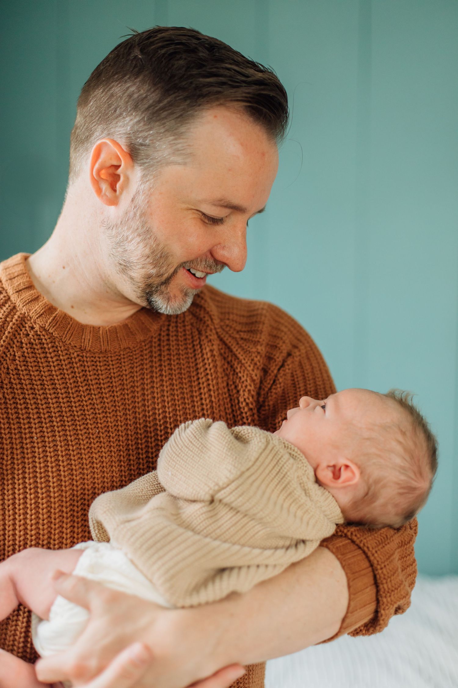 A bearded father in a rust sweater smiles warmly at his newborn baby he holds in his arms.