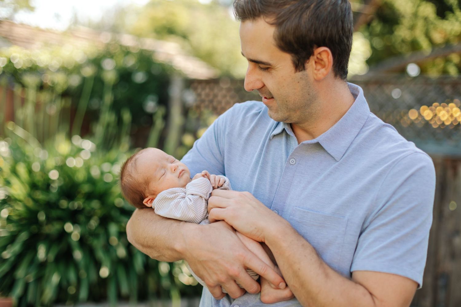 Father in light blue polo shirt tenderly cradling sleeping newborn baby outdoors in garden setting.