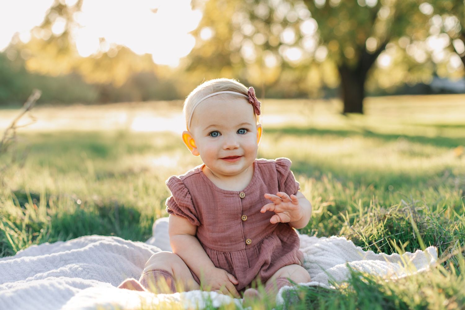 An adorable baby girl in a pink romper sits on a blanket smiling in a sunny park.