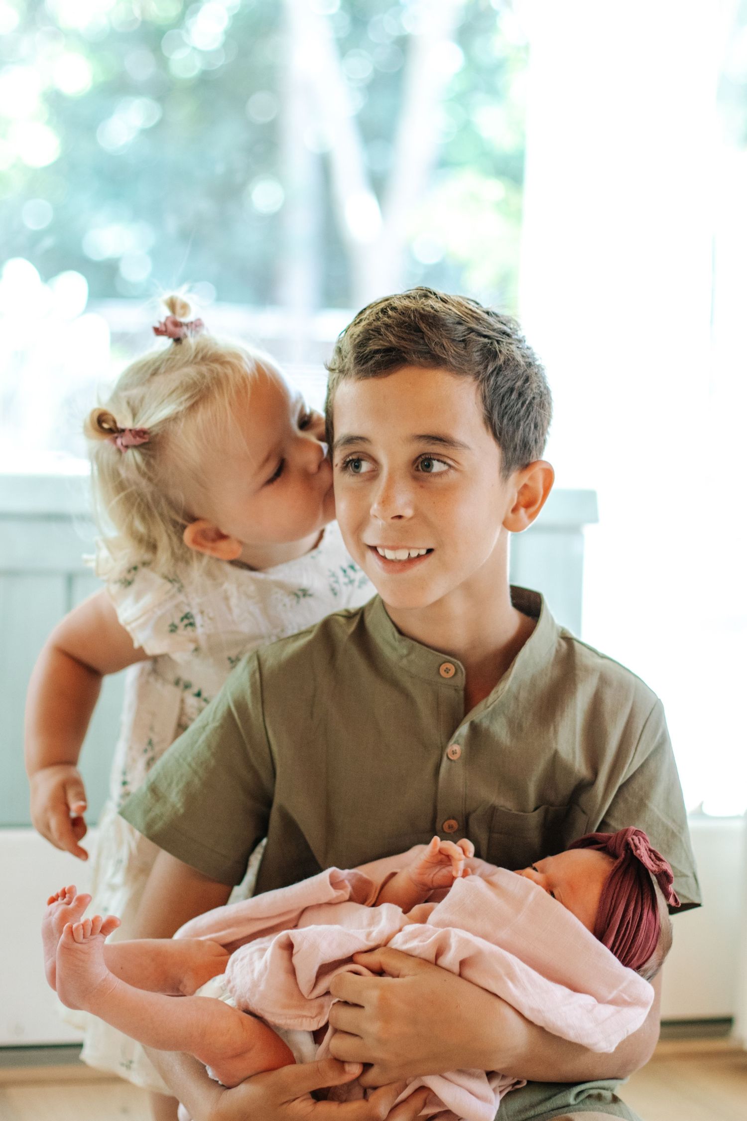 A smiling boy holds a newborn baby in pink while a toddler girl gives him a sweet kiss on the cheek.